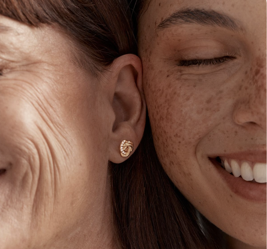 Close-up of two smiling women with freckles, one showing her ear with a gold twisted link earring with a small embedded diamond.