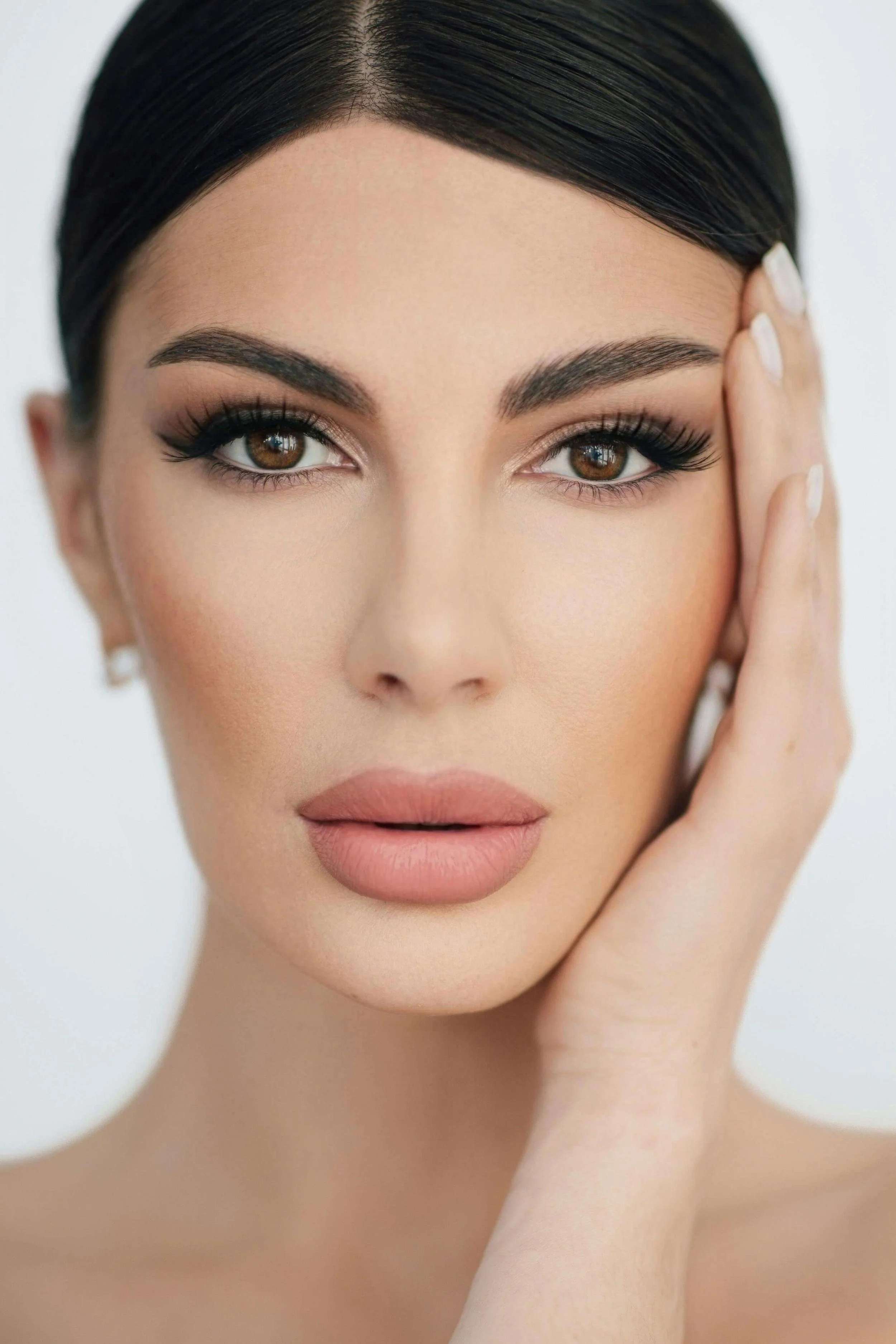 Close-up portrait of a woman with styled dark hair, brown eyes, and makeup including eyeshadow, eyeliner, and nude lipstick. She is touching her face gently with her right hand.