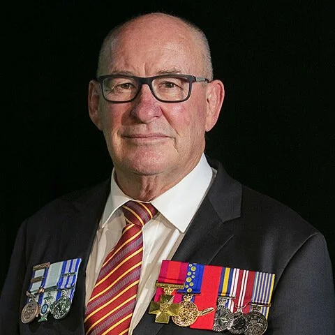 Portrait of an elderly man wearing glasses, a dark suit, a white shirt, and a striped red and gold tie, with military medals and ribbons on his chest.