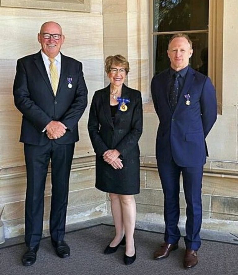 Three people standing indoors on a carpeted floor, wearing formal attire and medals, smiling at the camera.