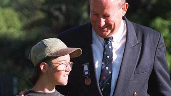 A man in a suit with medals on his lapel smiling and talking to a boy wearing glasses, a baseball cap, and a hoodie, outside with greenery in the background.
