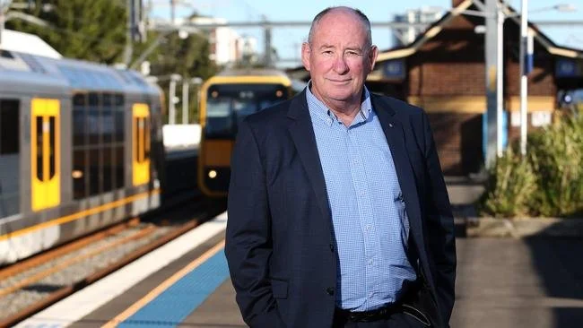 A man in a suit standing on a train platform with trains and tracks in the background.