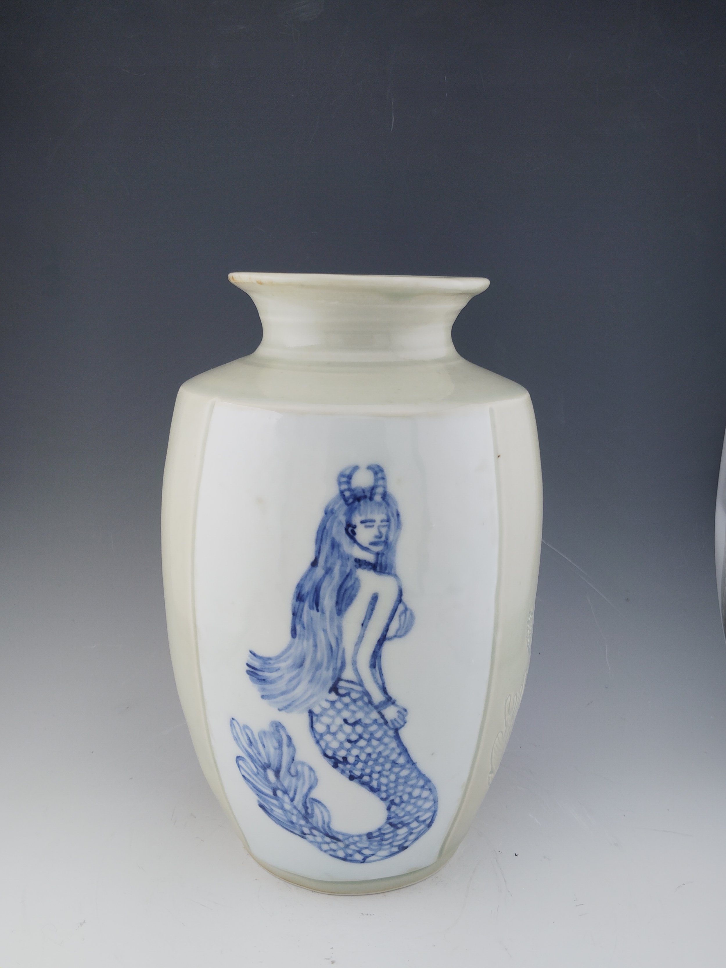 Ceramic vase with blue illustration of a mermaid with long hair and horns on a dark background.