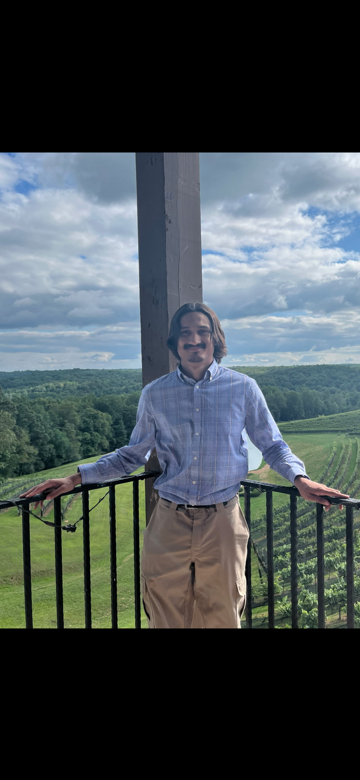A man standing on a balcony overlooking a lush, green landscape with vineyards and trees, under a partly cloudy sky.
