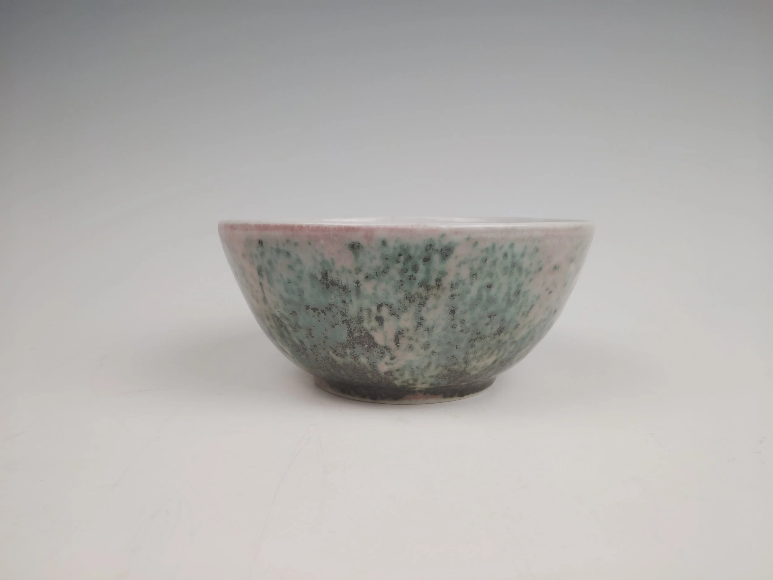 A ceramic bowl with a speckled glaze, predominantly green and pinkish hues, against a plain white background.