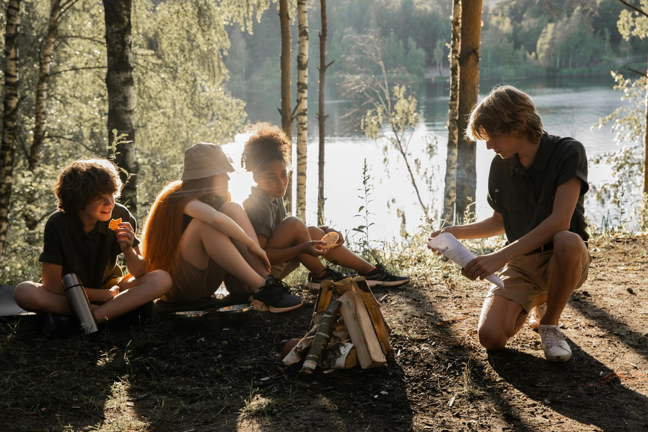 A group of five children sitting on the ground near a campfire by a lake in a forest, with some holding food and others looking at a map, enjoying a camping trip in nature during sunset.