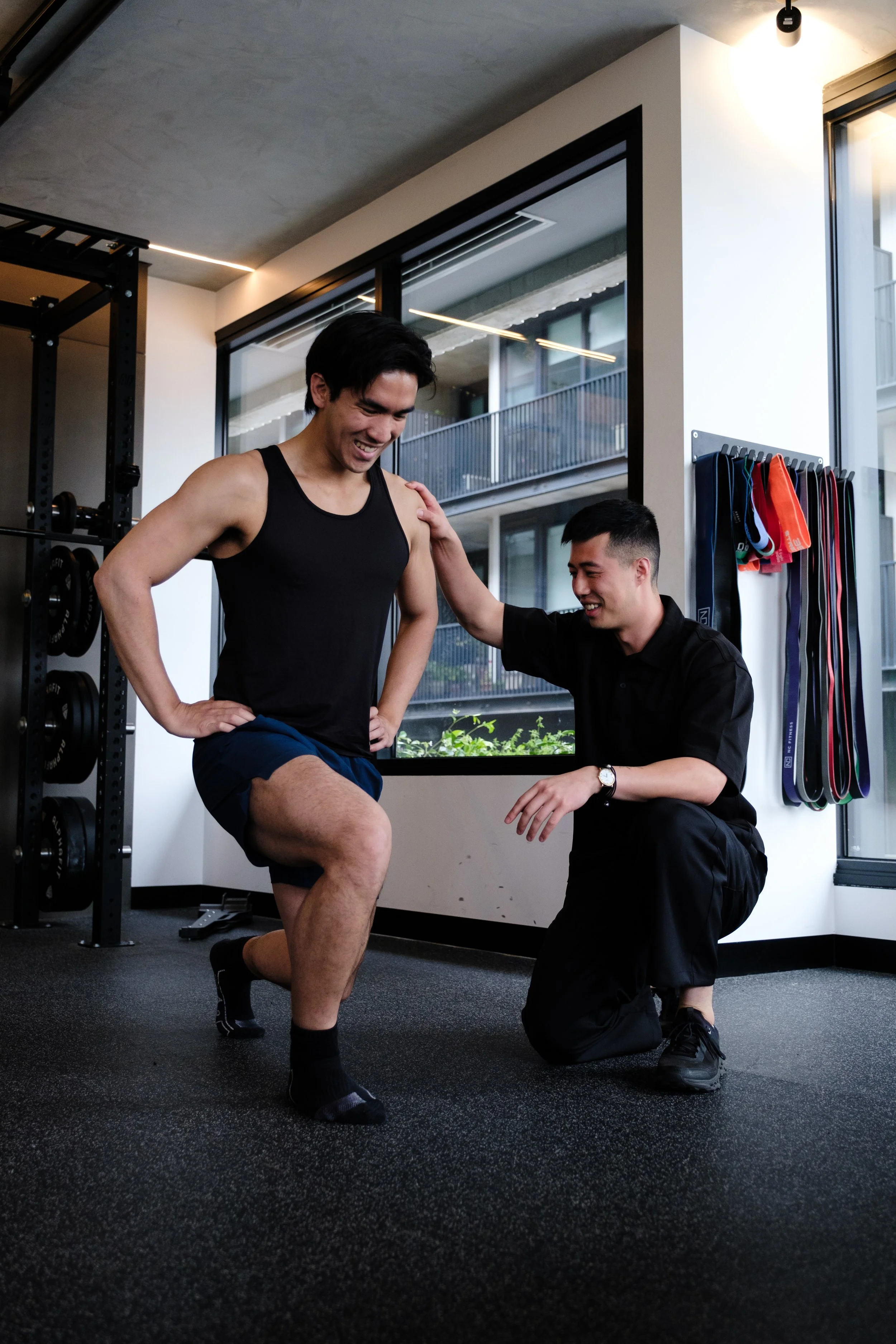 A man in a black tank top and shorts is performing a knee exercise with assistance from a trainer who is kneeling beside him in a gym.