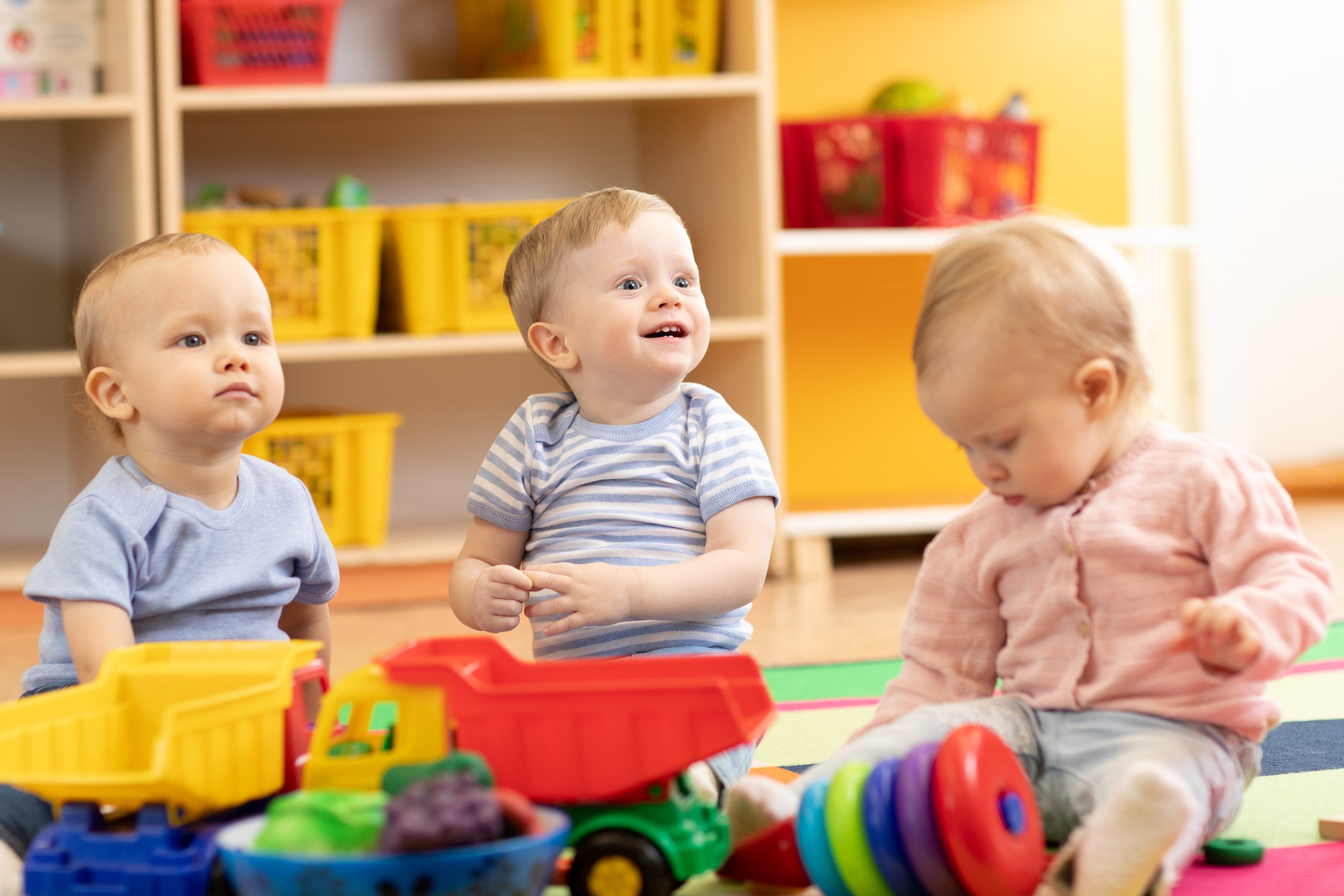 Three young children playing with toys on a colorful rug in a playroom with shelves and storage bins behind them.