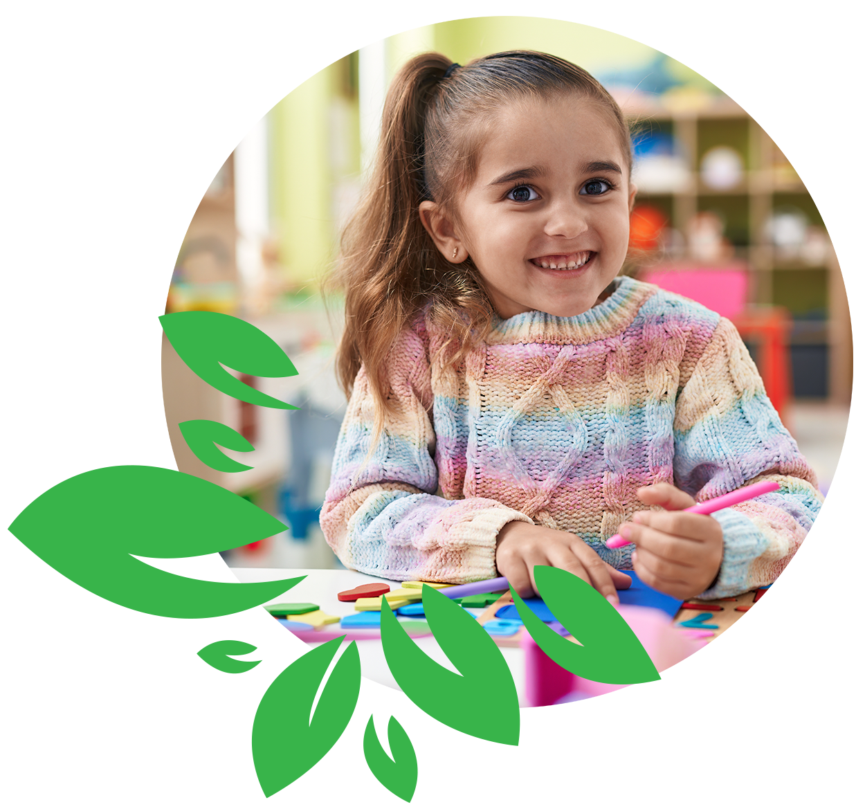 A young girl with long, light brown hair in a ponytail, smiling while sitting at a table with colorful craft supplies, in a classroom setting.