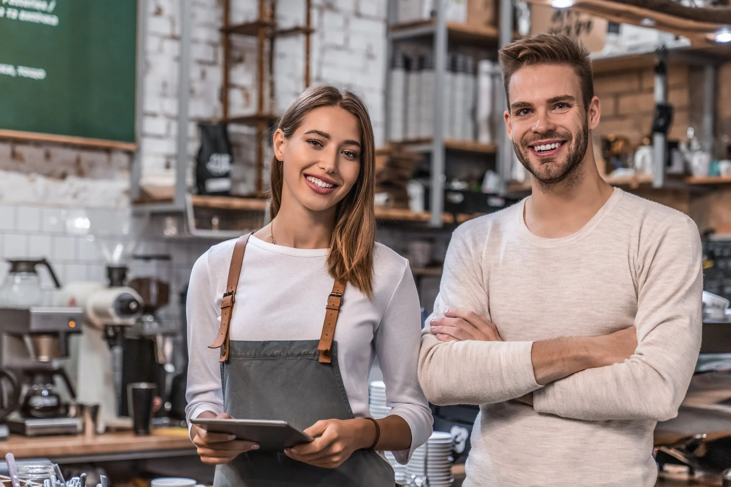 To smilende personer i en kafé eller restaurant, med hyller og kjøkkenutstyr i bakgrunnen.