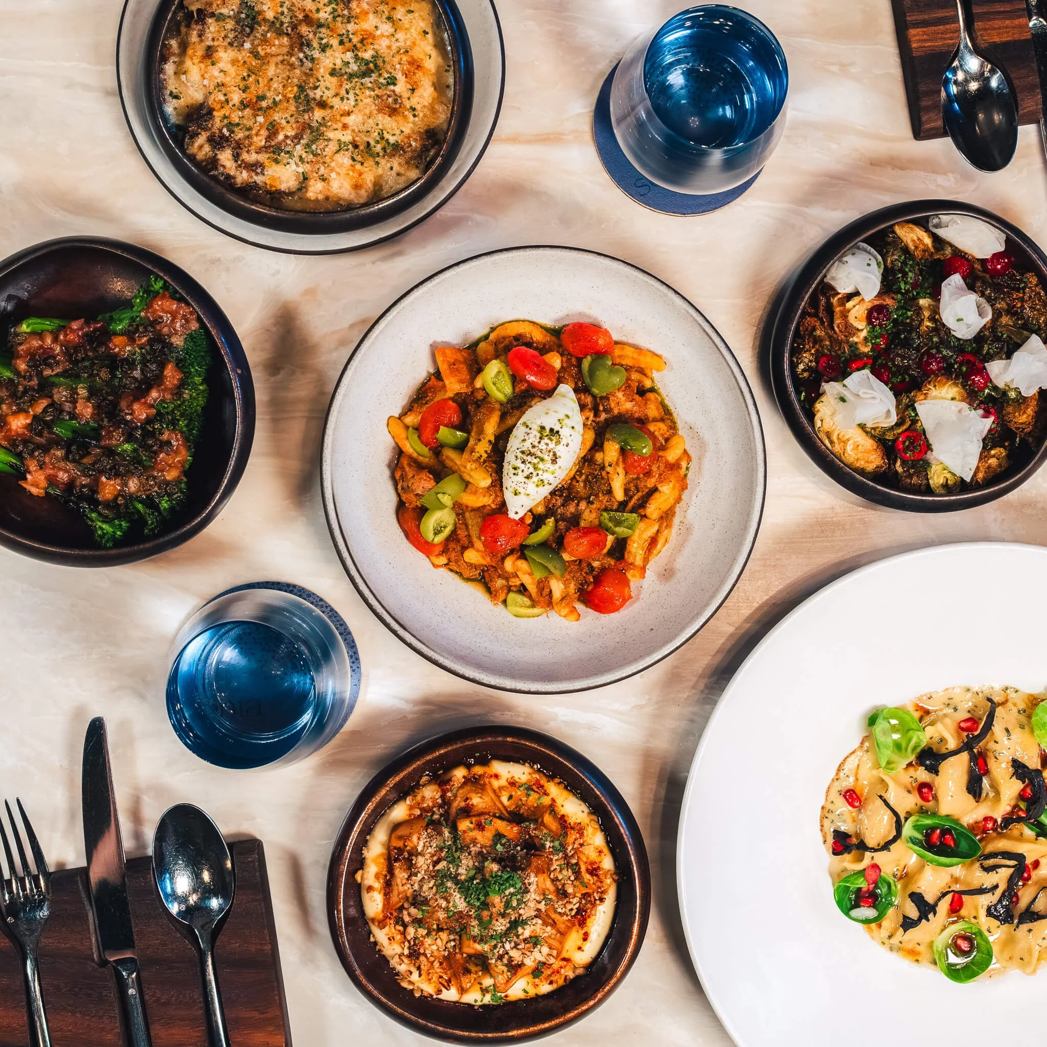 Top-down view of a table with various dishes, including a pasta with cherry peppers on a white plate, a pasta with herbs and cheese in a dark bowl, roasted vegetables topped with herbs, a tomato salad with white cheese, a baked dish with melted cheese and breadcrumbs, another baked dish with cheese and vegetables, and two glasses of blue-tinted water.