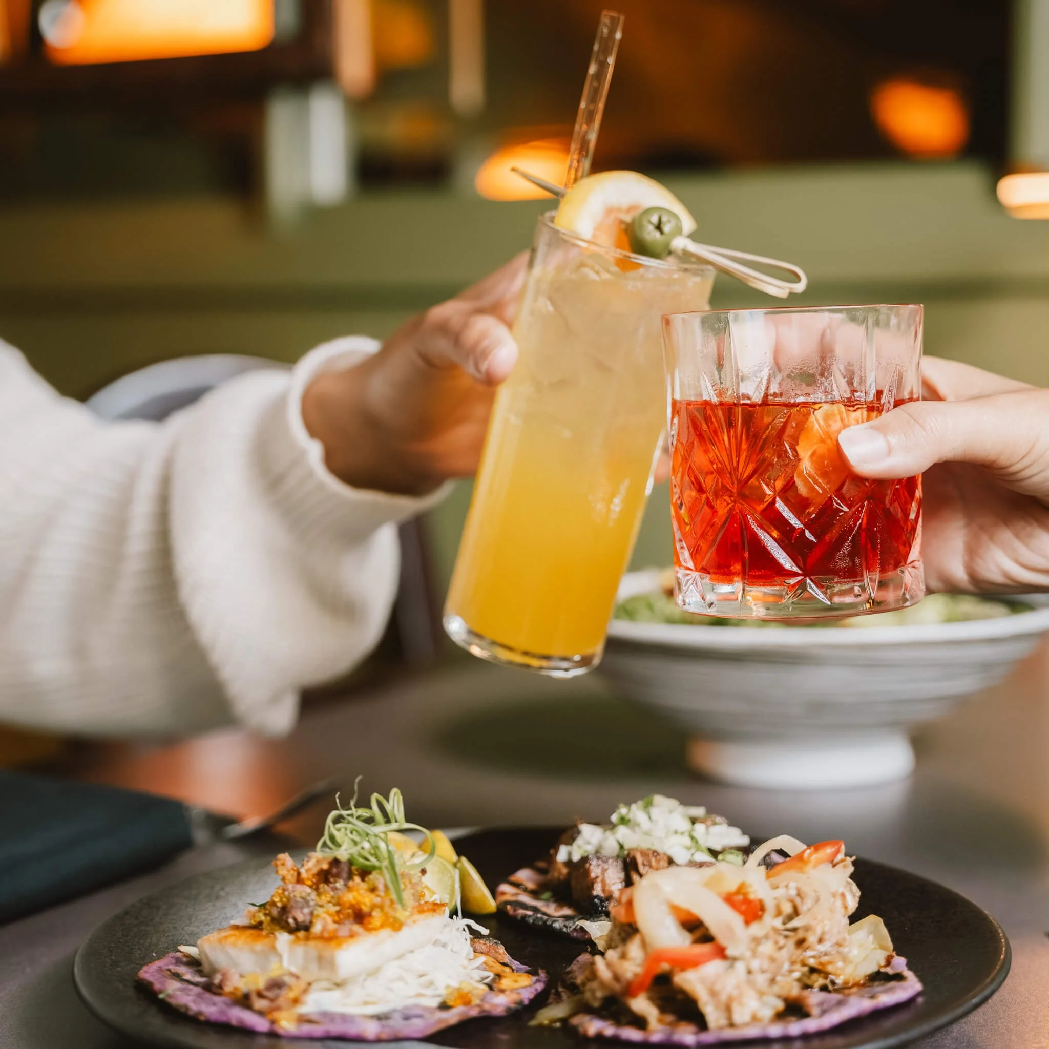 Two people clinking glasses of colorful cocktails over a table with various brightly decorated appetizers.