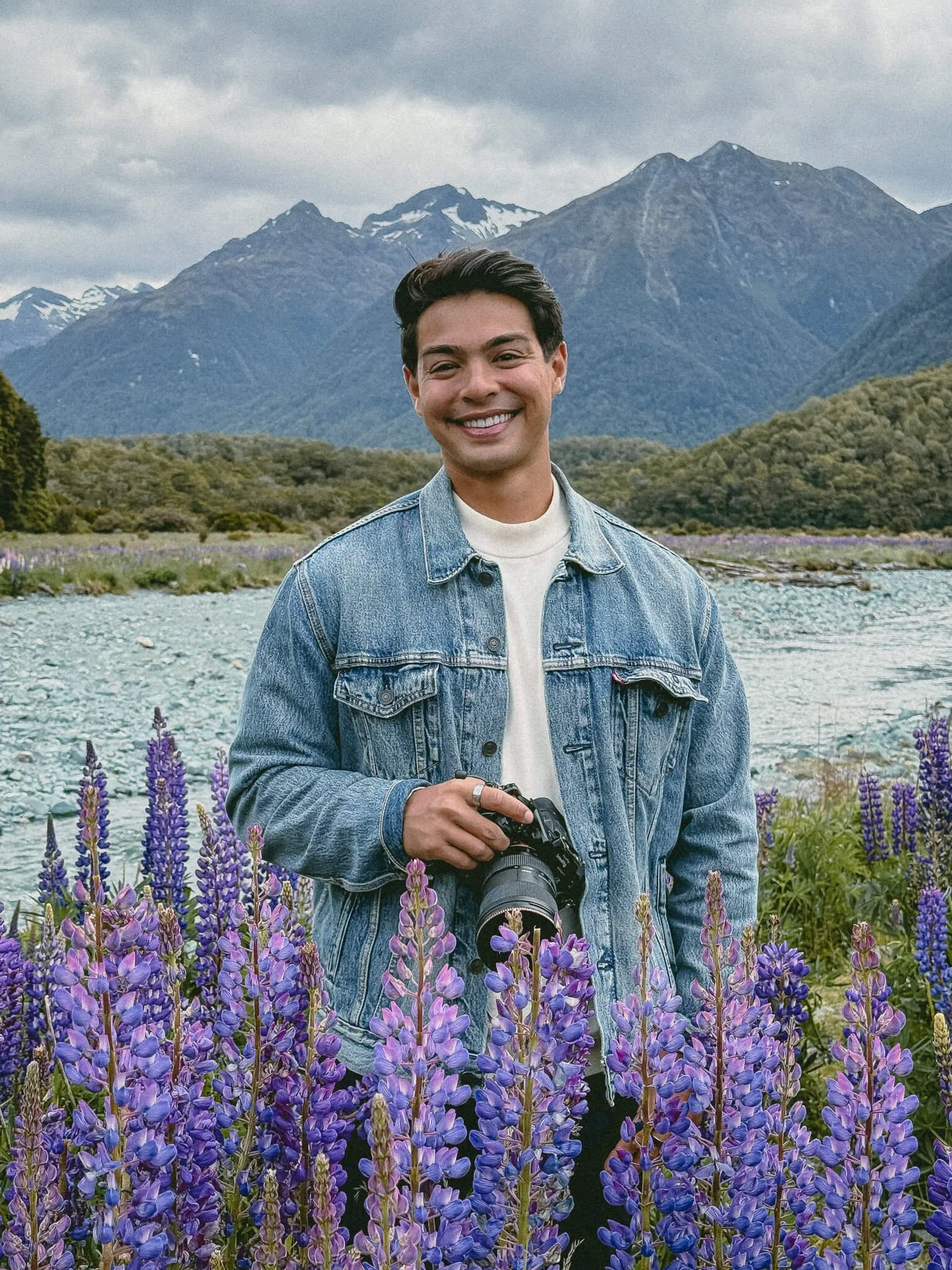 A smiling man holding a camera in a field of purple flowers with mountains and a cloudy sky in the background.
