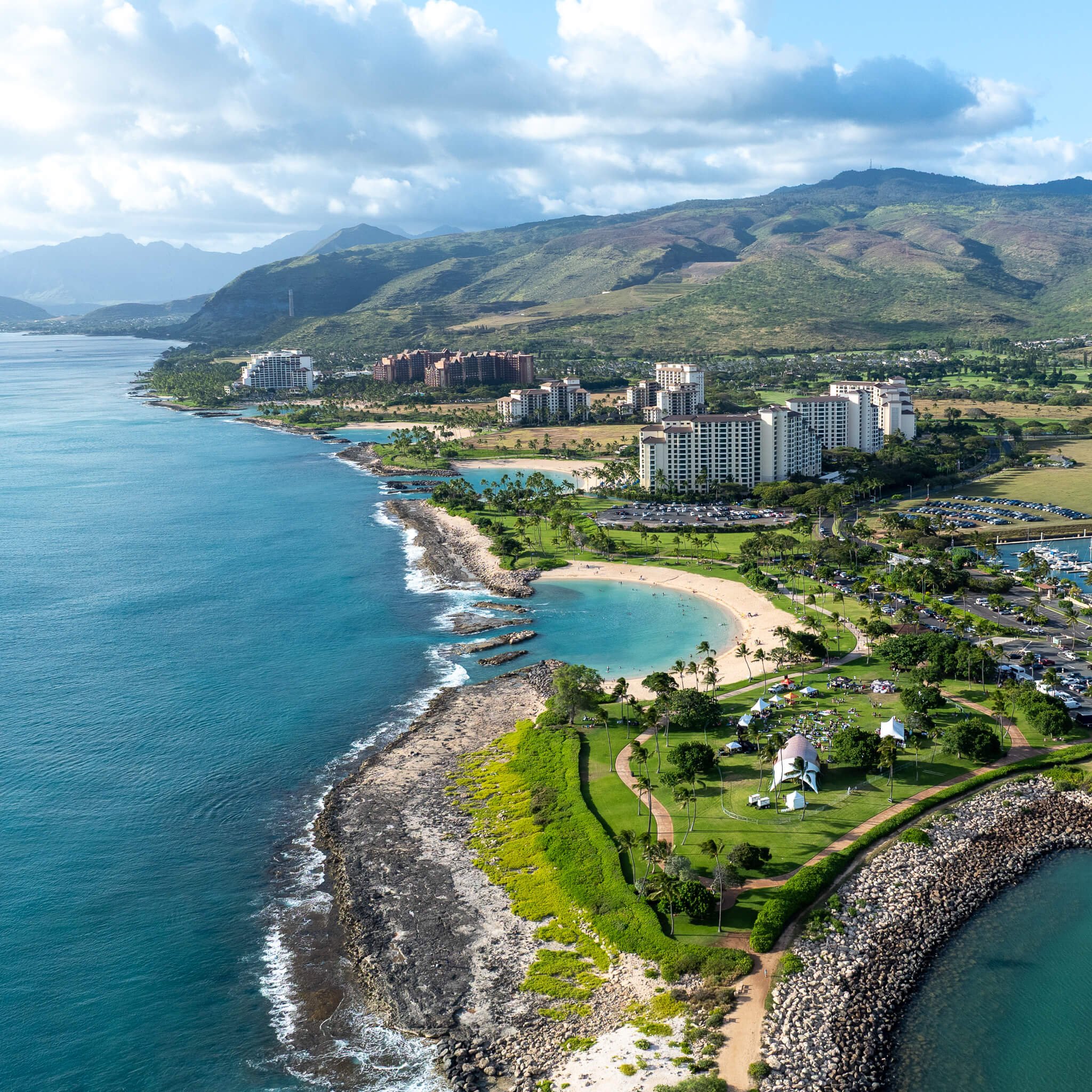 Aerial view of a coastal city with a beach, high-rise buildings, marina, green parks, and mountainous backdrop.