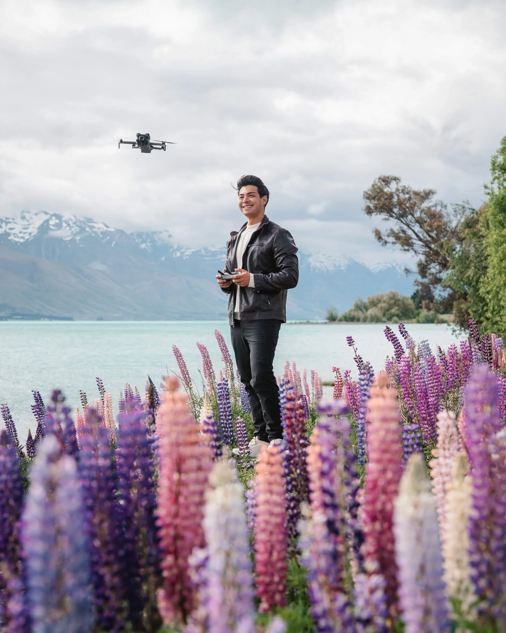 A man holding a drone remote controller, standing among colorful flowers at a lakeside with mountains in the background.