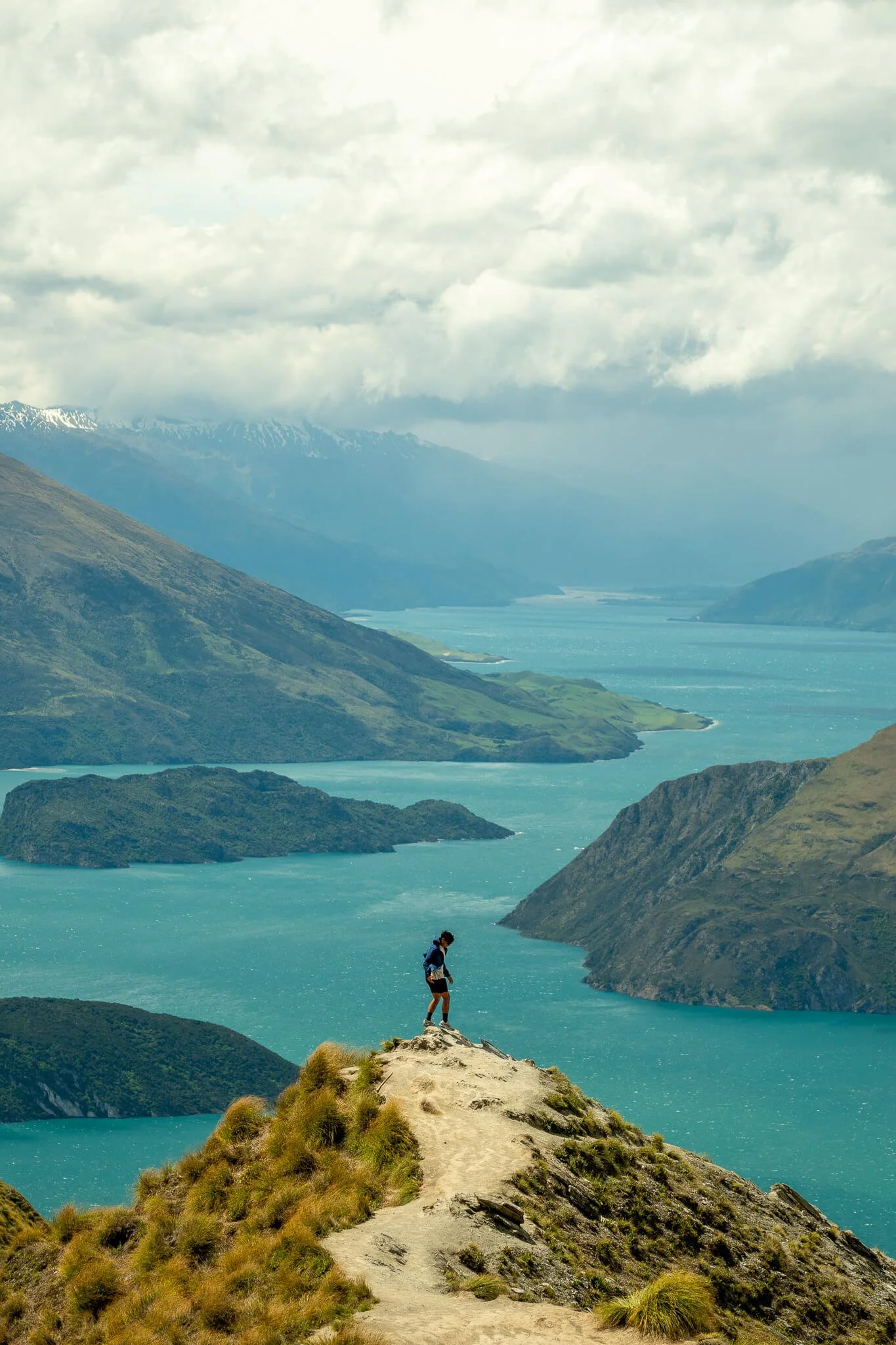 Hiker standing on a trail on a mountain ridge overlooking a turquoise lake and green mountains, with snow-capped peaks in the background under cloudy sky.
