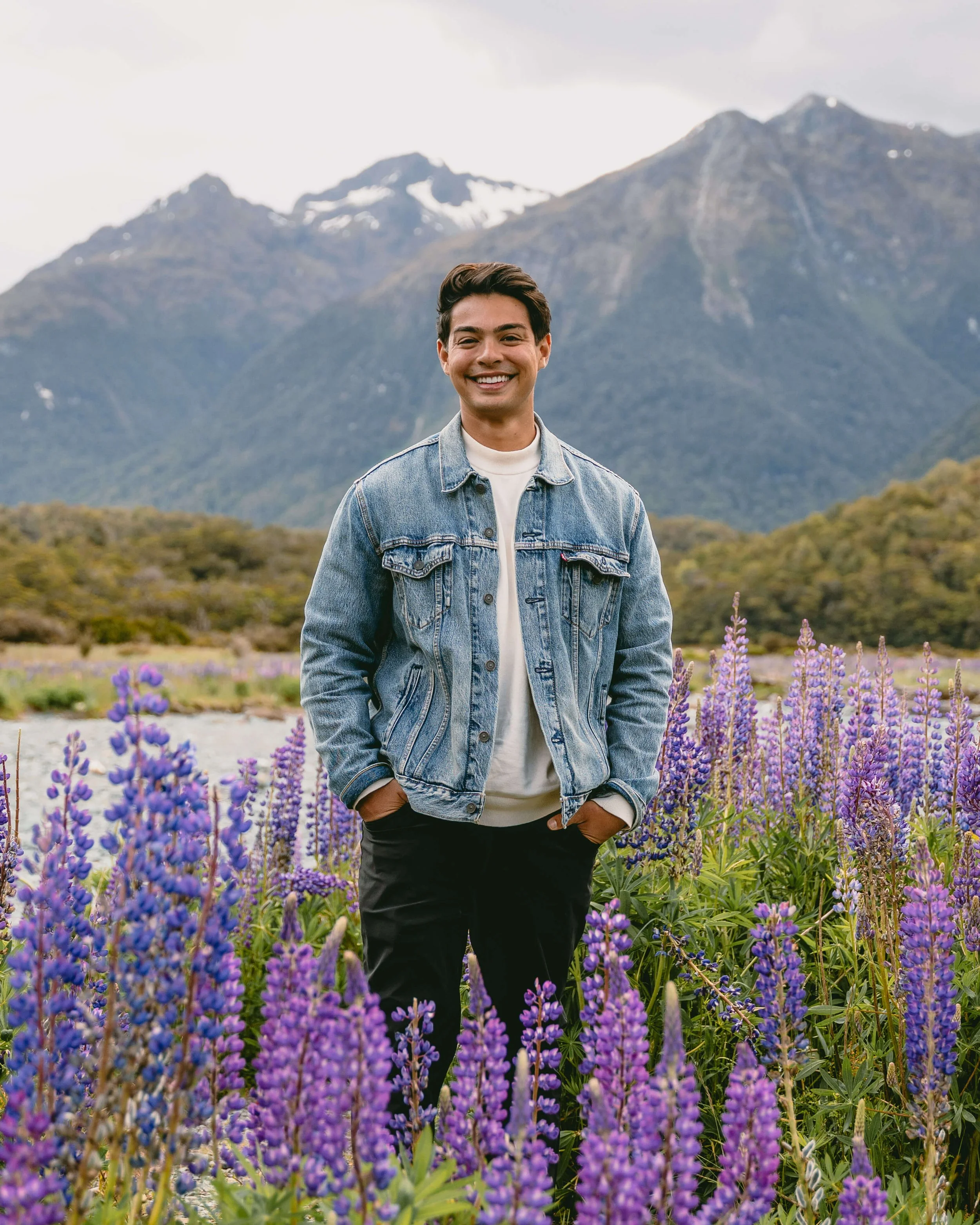 A smiling young man in a denim jacket standing in a field of purple flowers with mountains in the background.