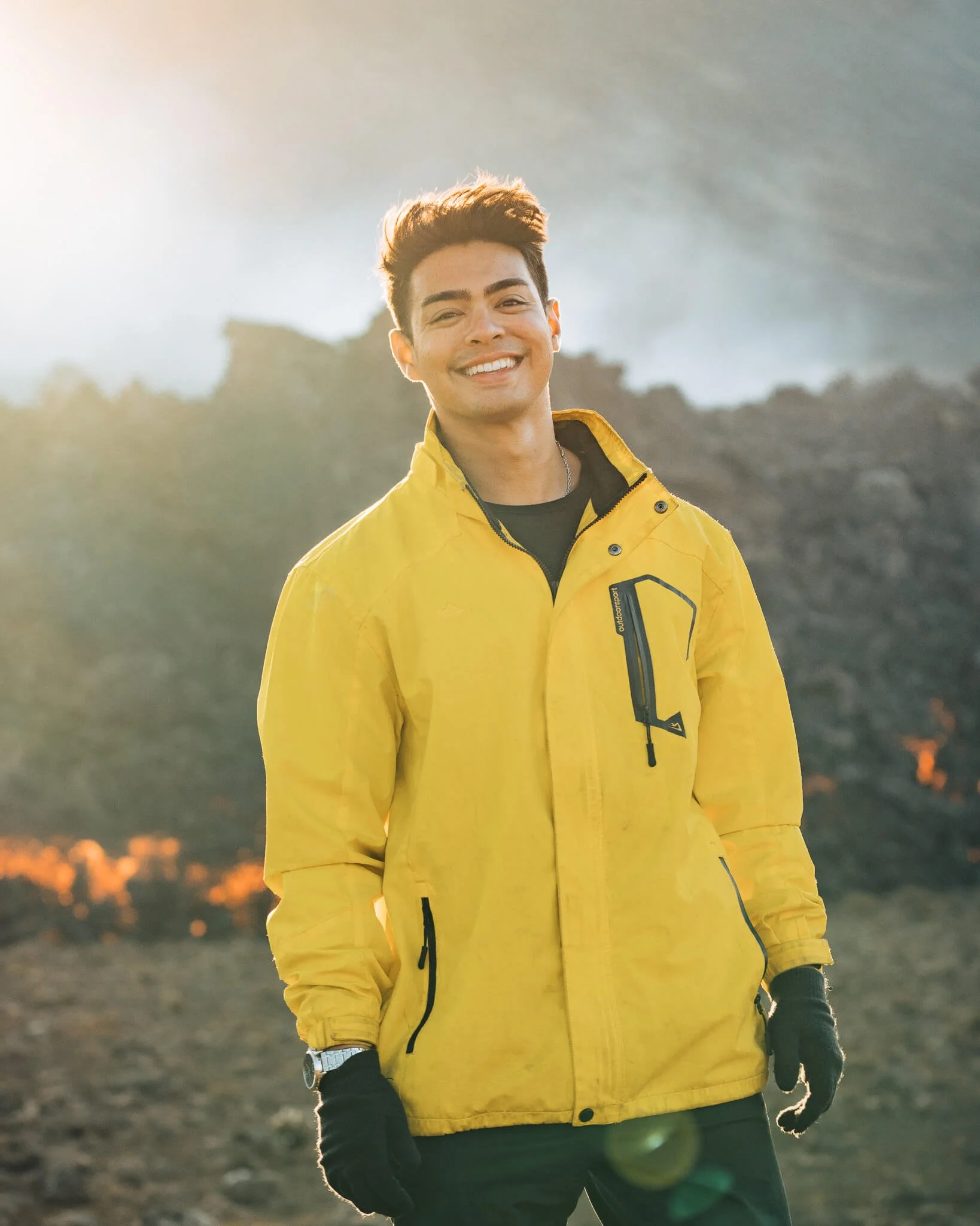 Young man outdoors wearing a yellow jacket and black gloves, smiling at the camera, with mountains and sunlight in the background.