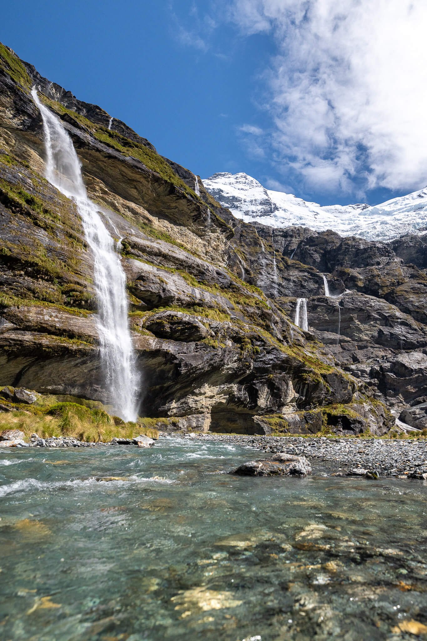 Mountain landscape with waterfalls, snow-capped peaks, and a flowing river.