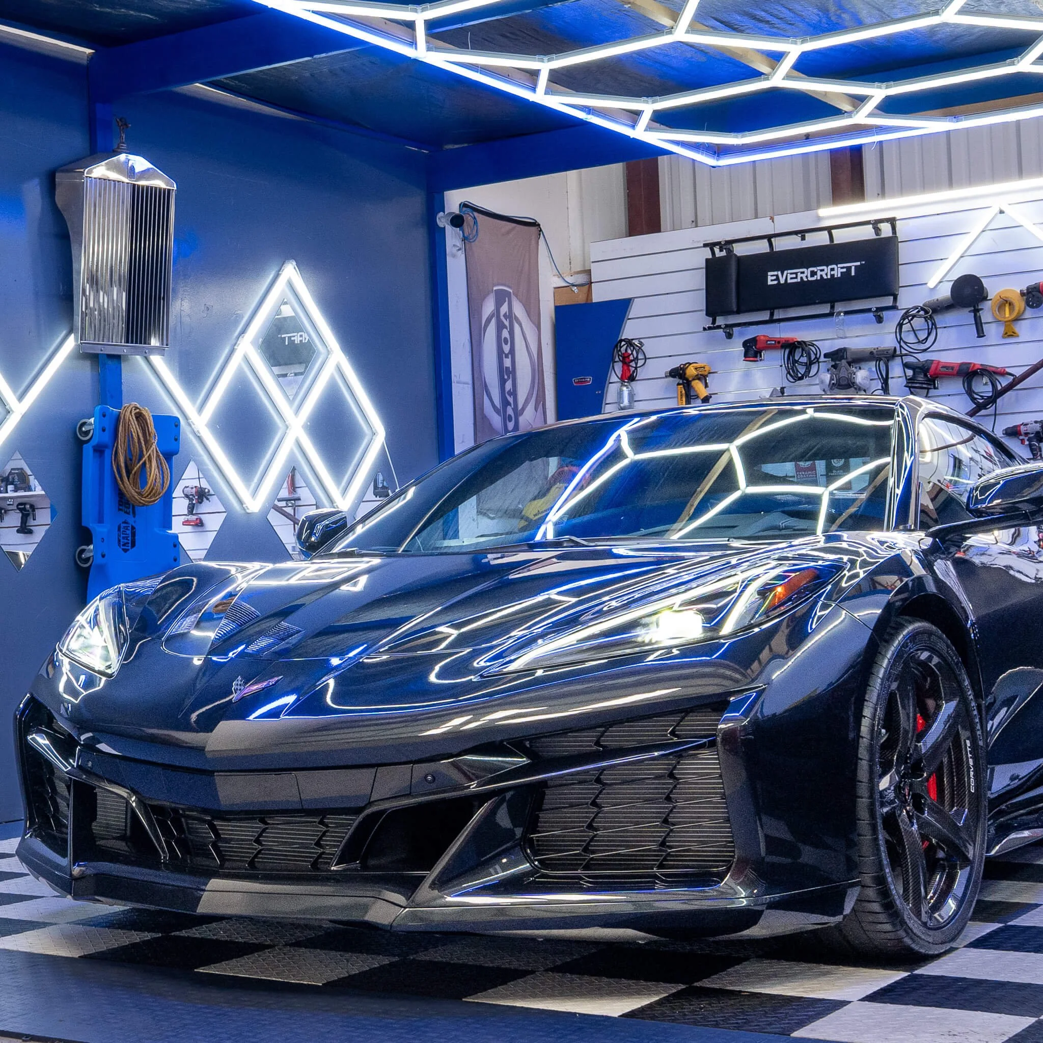 A black sports car parked inside a garage with neon blue and white lighting and various tools hanging on a white wall.
