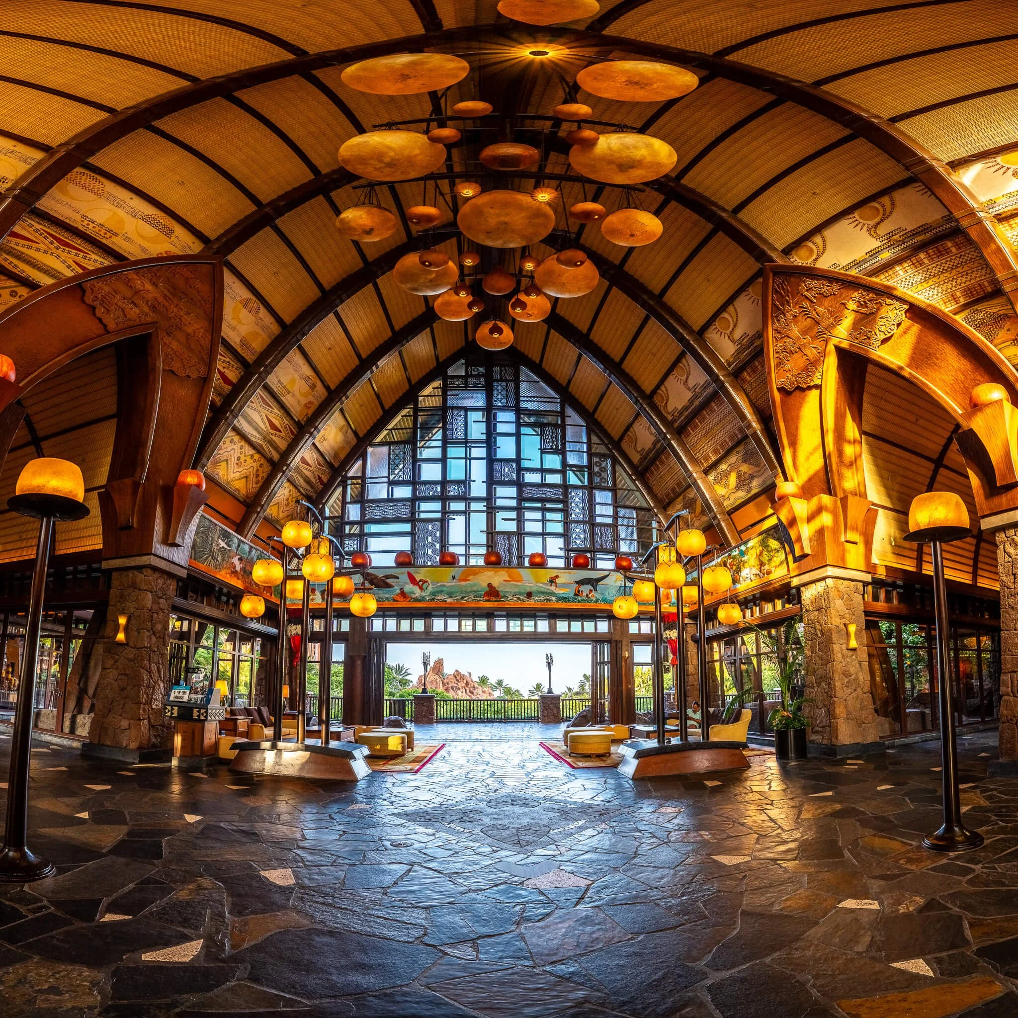 Interior of a large, ornate building with high, arched ceiling, wooden accents, and large windows, decorated with hanging lamps and artwork, overlooking a balcony with outdoor view.