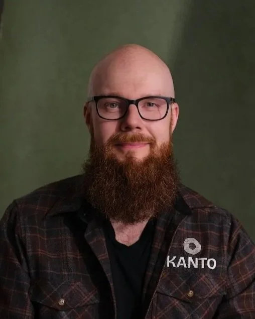 Bald man with glasses and a full beard, wearing a plaid shirt with a logo that reads 'KANTO' on the chest, smiling against a green background.