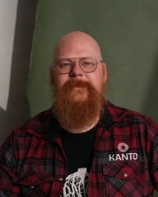 A man with glasses and a beard wearing a red and black plaid shirt and a black T-shirt with white text, standing in front of a green backdrop.