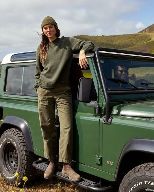 Mujer con suéter y pantalones cargo posa con un vehículo verde en un paisaje campestre.