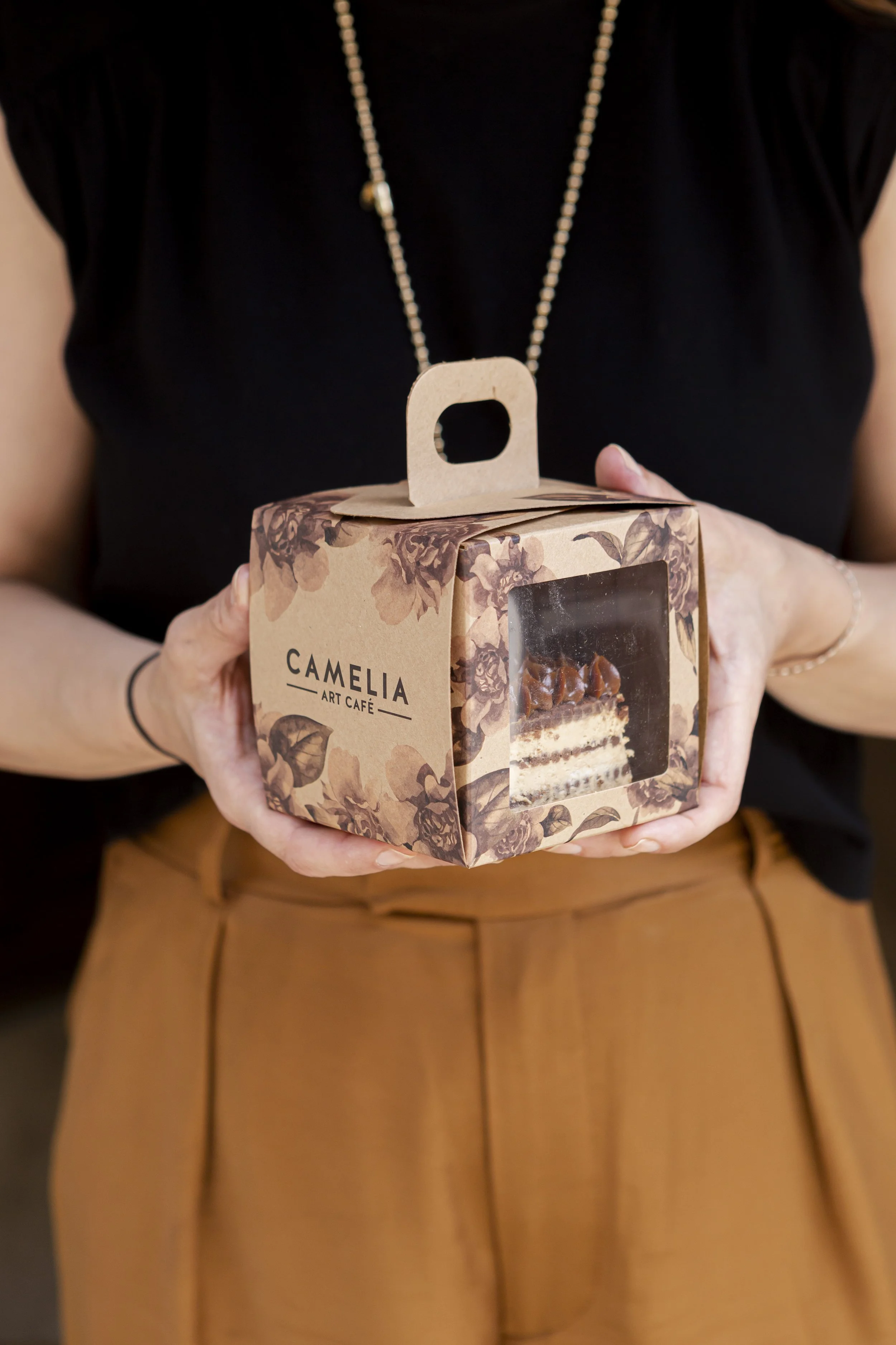 Persona sosteniendo caja de pastel con diseño floral y ventana de acrílico de la marca Camelia Art Café.