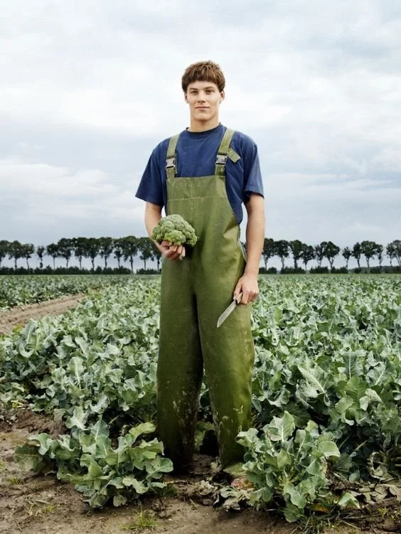 Joven en campo agrícola sosteniendo un brócoli y una cuchilla, vestido con overol verde y camiseta azul, con árboles en el fondo y cielo nublado.