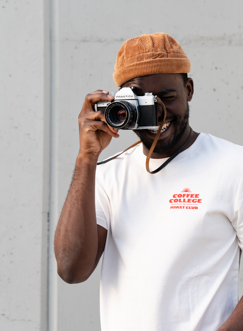 Joven sonriente con gorro marrón y camiseta blanca que sostiene una cámara vintage frente a un fondo gris.