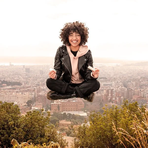 Una mujer con cabello rizado y sonrisa sentada en posición de meditación en un paisaje urbano, con un cielo despejado y árboles en primer plano.