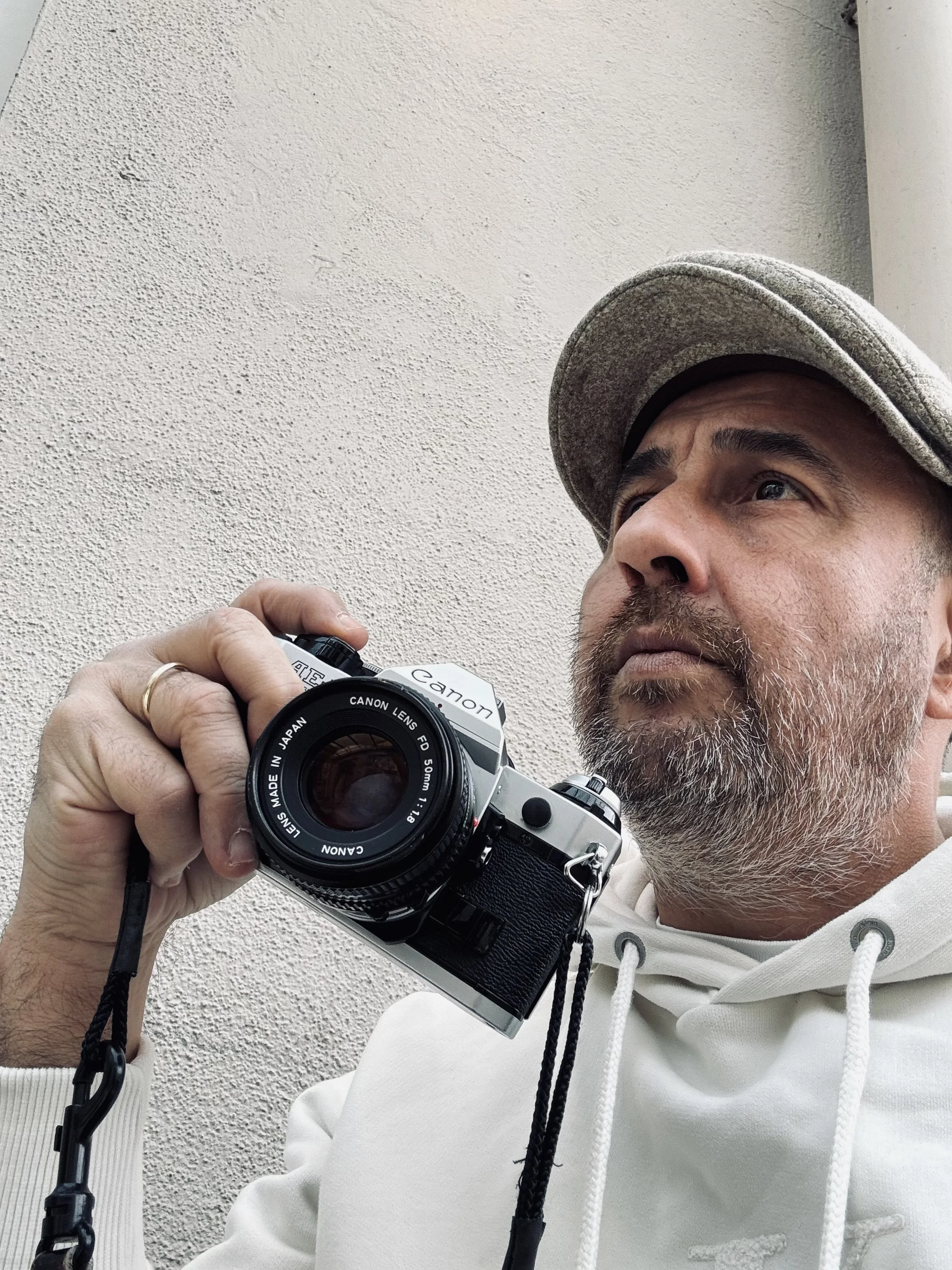 Ein Mann trägt einen grauen Hut und eine weiße Kapuzenjacke, hält eine Kamera mit einem Objektiv und schaut nach oben.