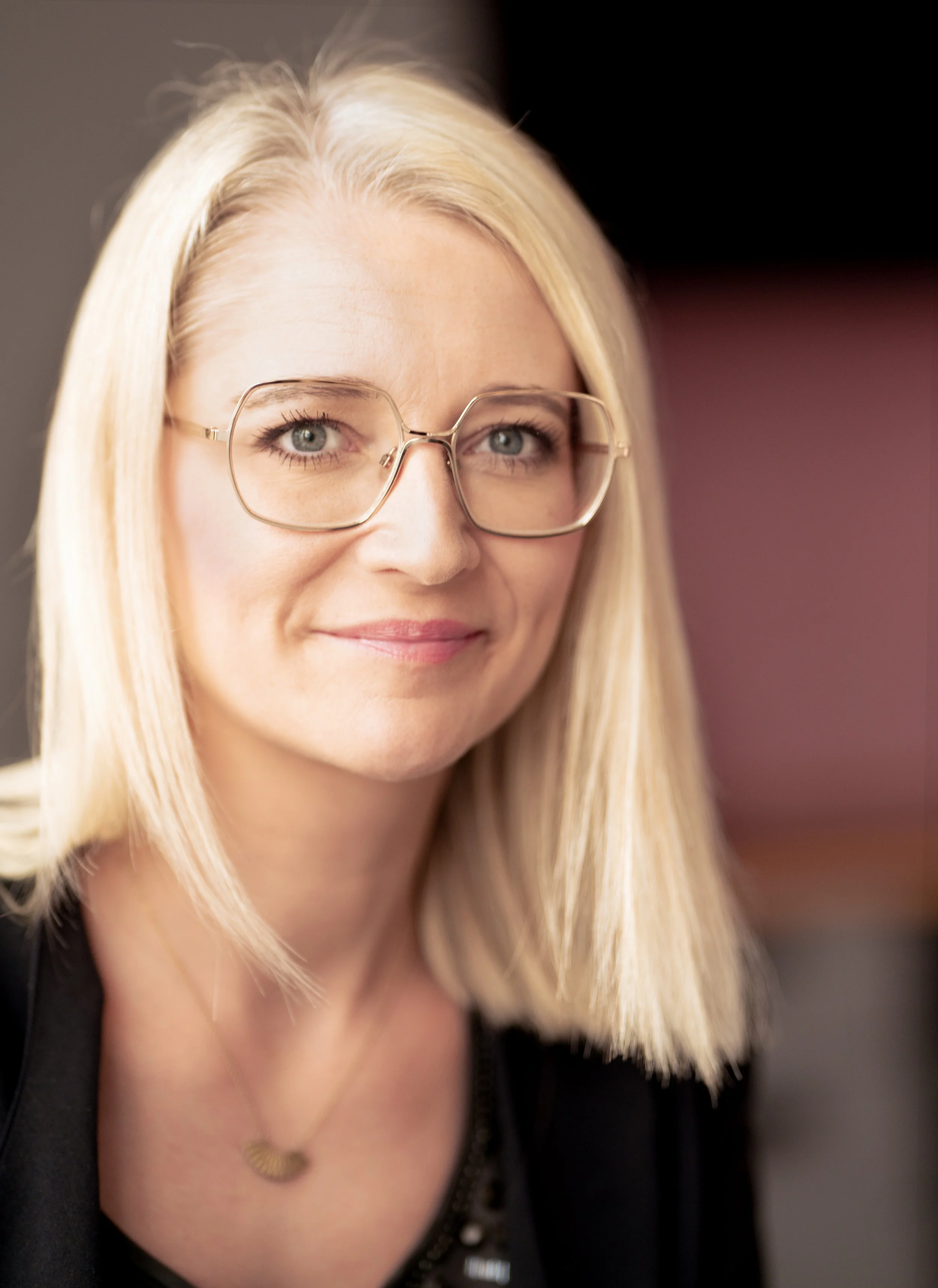 Portræt Rikke Lundsfryd, business wingwoman, med blond hår, briller og et smil, iført sort tøj og en guldhalskæde.