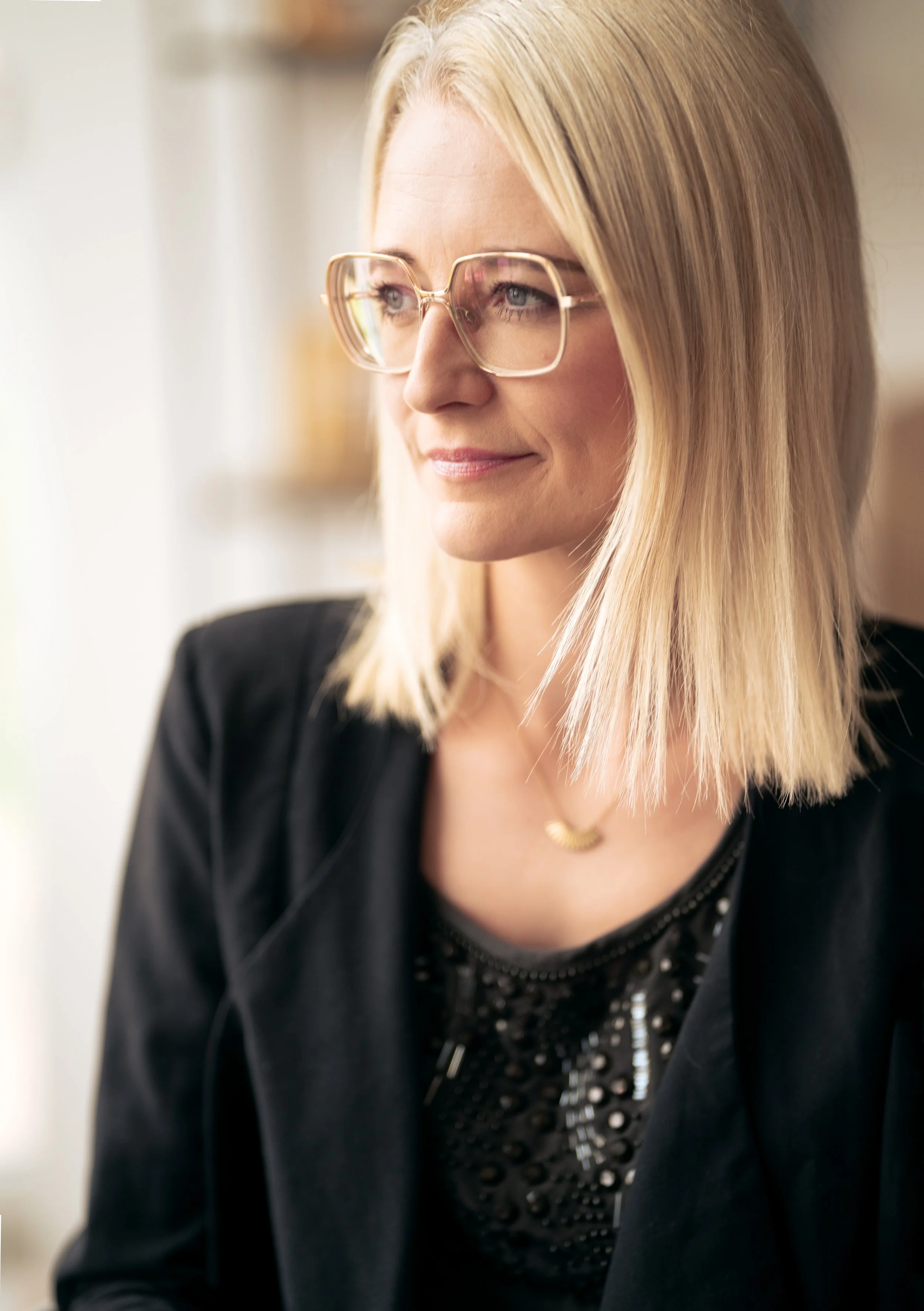 Rikke Lundsfryd, business wingwoman, kvinde med briller, der ser ud af et vindue, iført sort jakke og en sort bluse med perler.