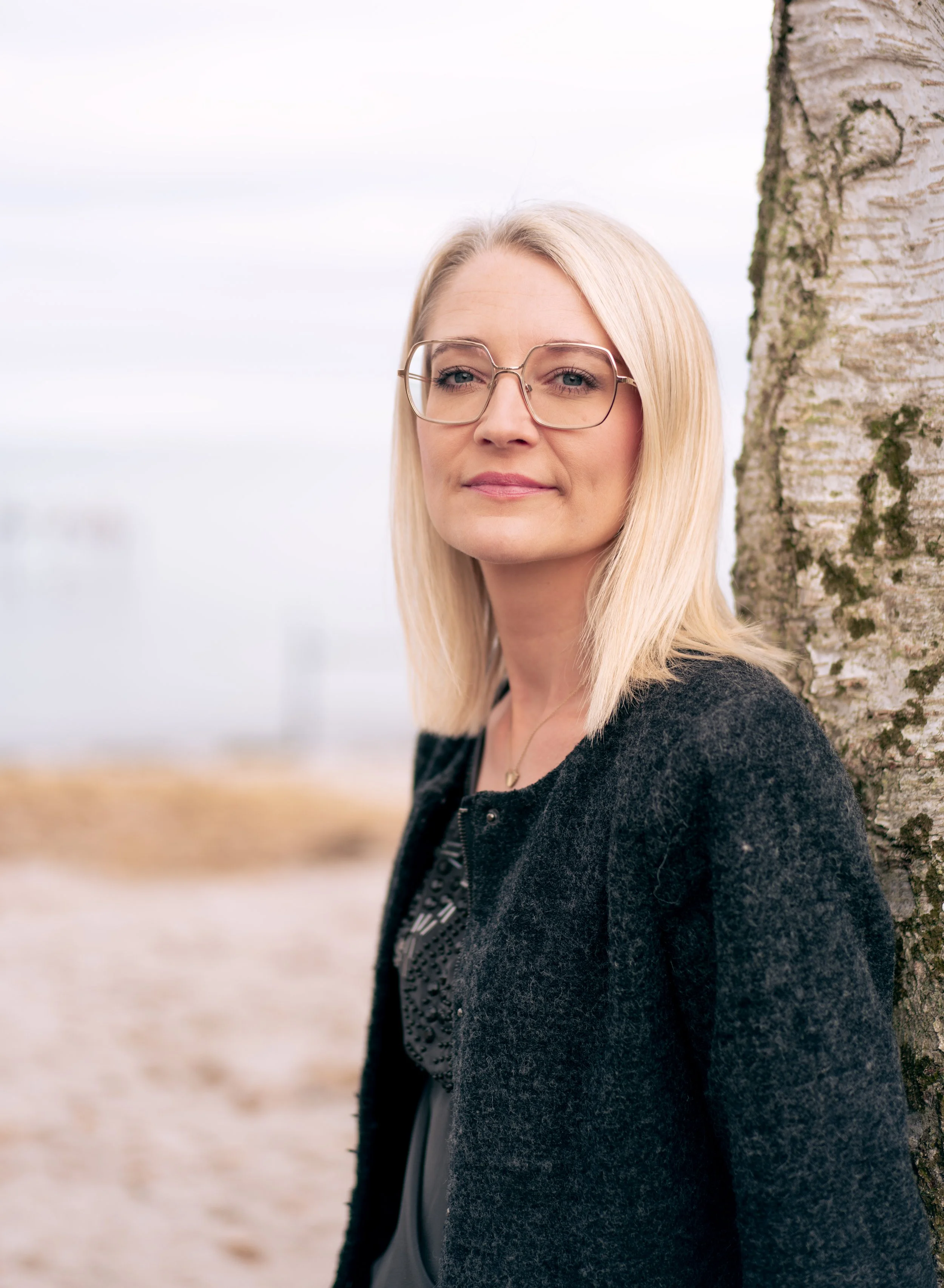 Rikke Lundsfryd, business wingwoman, en blond kvinde med briller står ved et birketræ udendørs med en sløret baggrund.