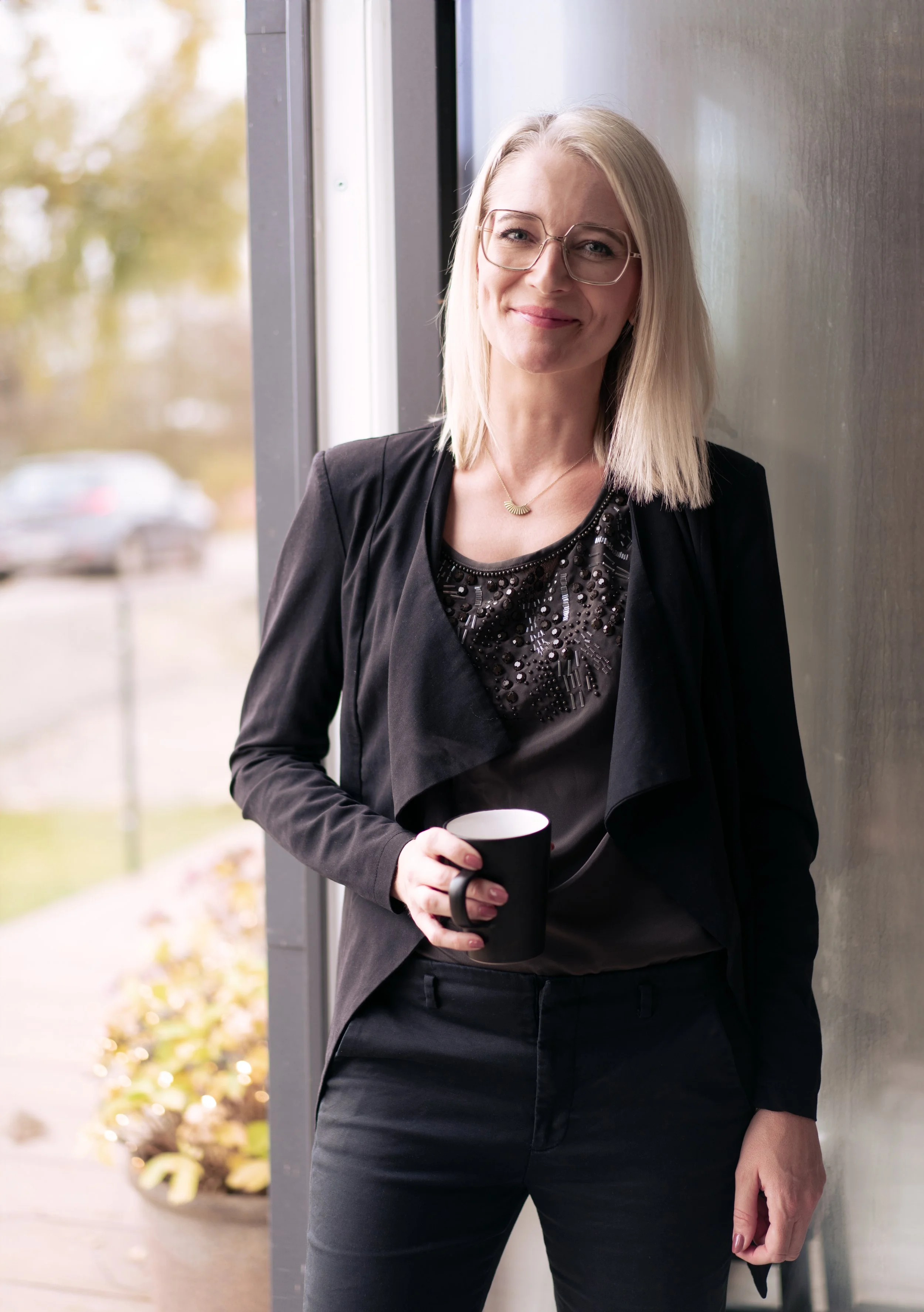 Rikke Lundsfryd, business wingwoman, har blond hår, iført sort tøj og briller, står ved et vindue og holder en sort kop.