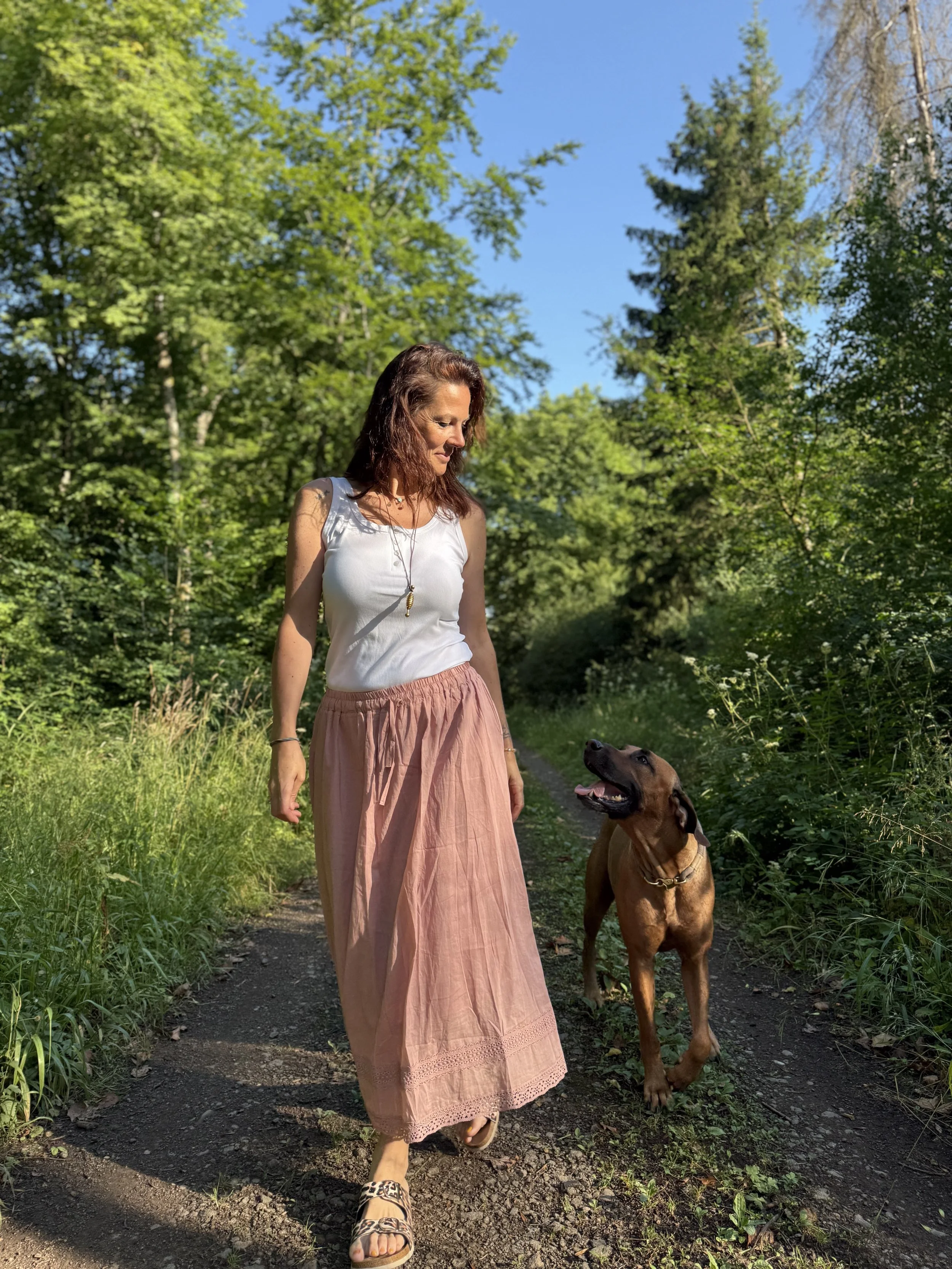 Eine Frau läuft mit einem Hund auf einem Waldweg. Es ist sonnig, und die Umgebung ist grün mit Bäumen und Gras.