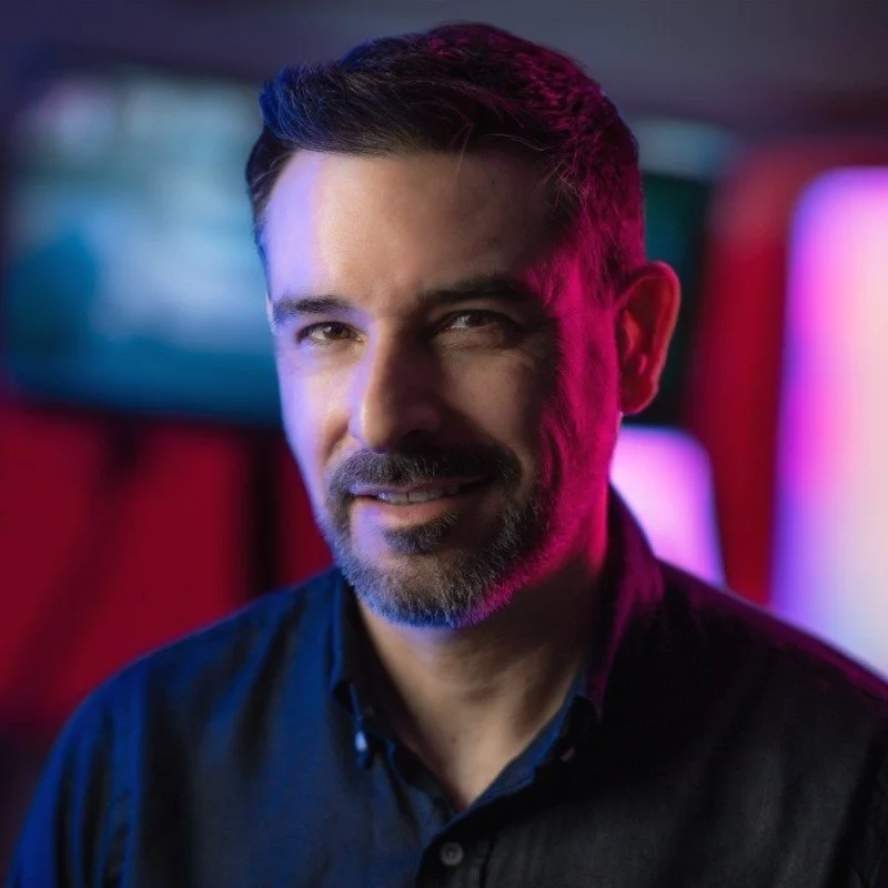 A man smiling, lit with colorful lighting, against a blurred background.