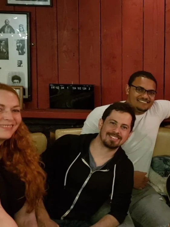 Group of three people smiling indoors, posing in a cozy setting with red wooden walls, framed pictures, a scoreboard, and comfortable seating.