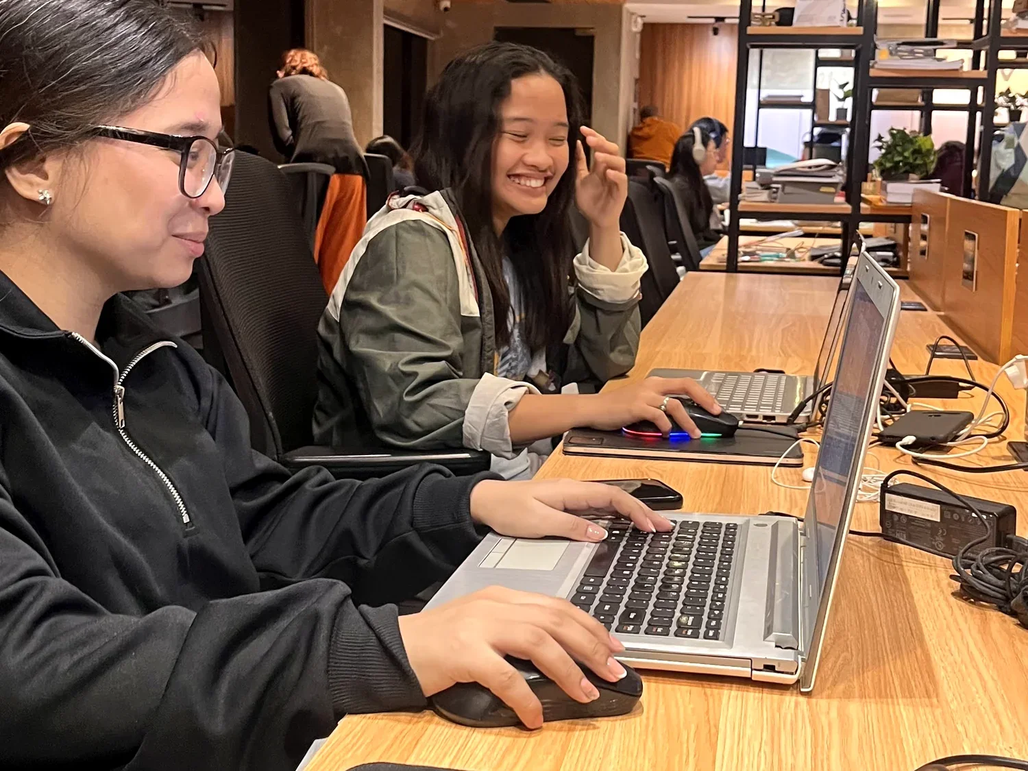 Two women sitting at a wooden desk working on laptops, one smiling and the other focused, in a shared workspace with other people in the background.