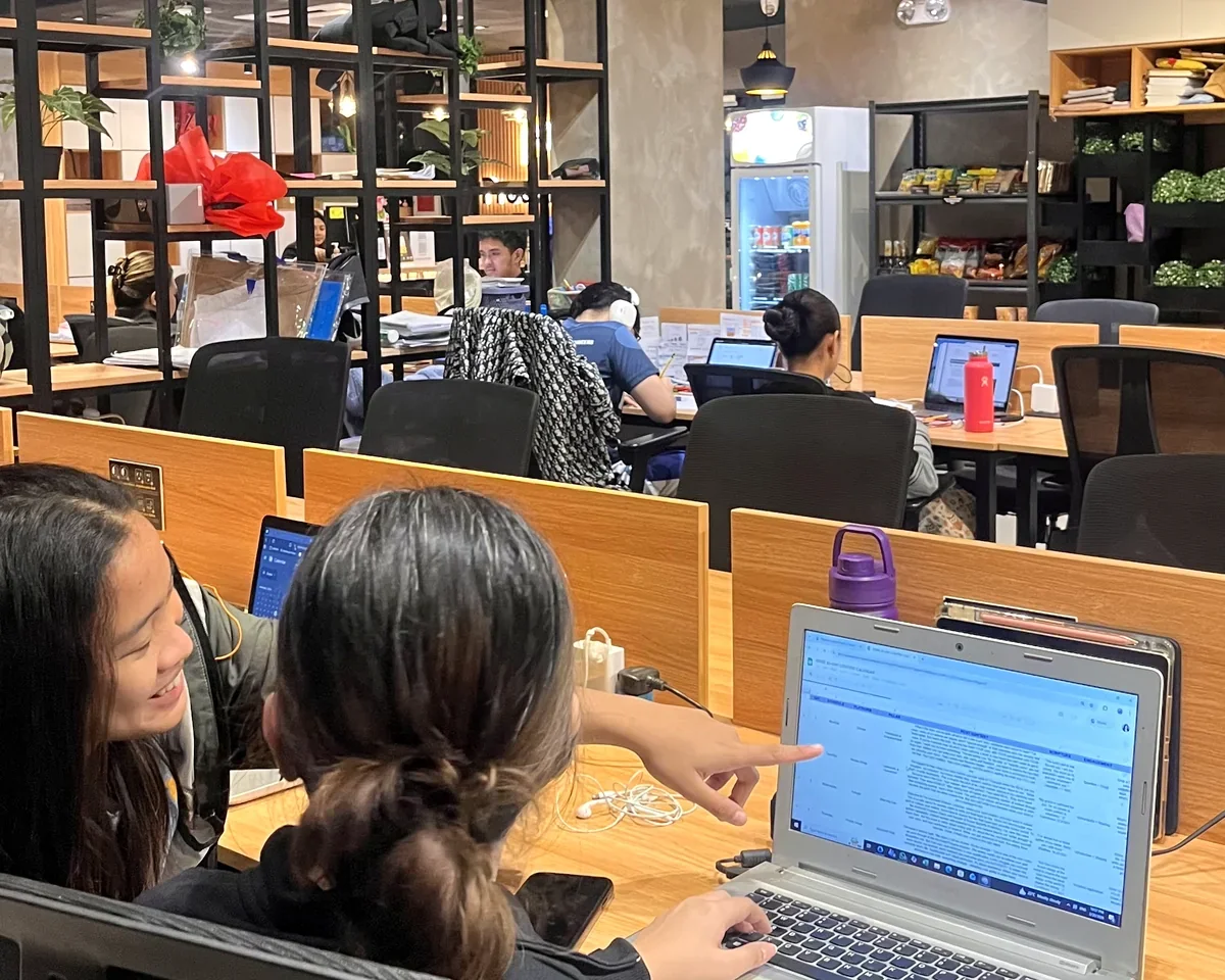 People working on laptops in a modern coworking space or cafe, with shelves, a refrigerator, and various personal and work items visible.