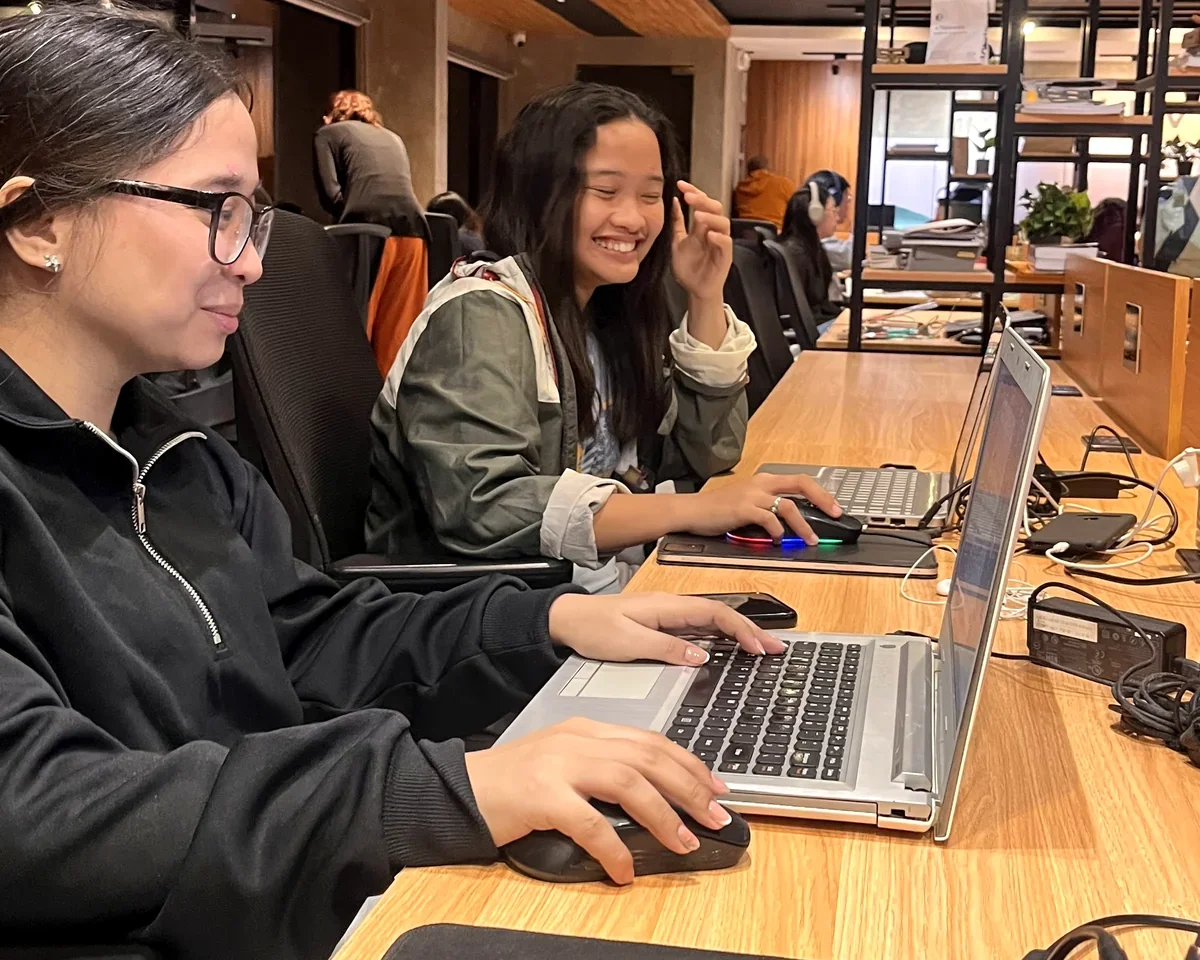 Two young women working on laptops at a long office desk in a modern workspace, smiling and focused on their screens, with other coworkers in the background.