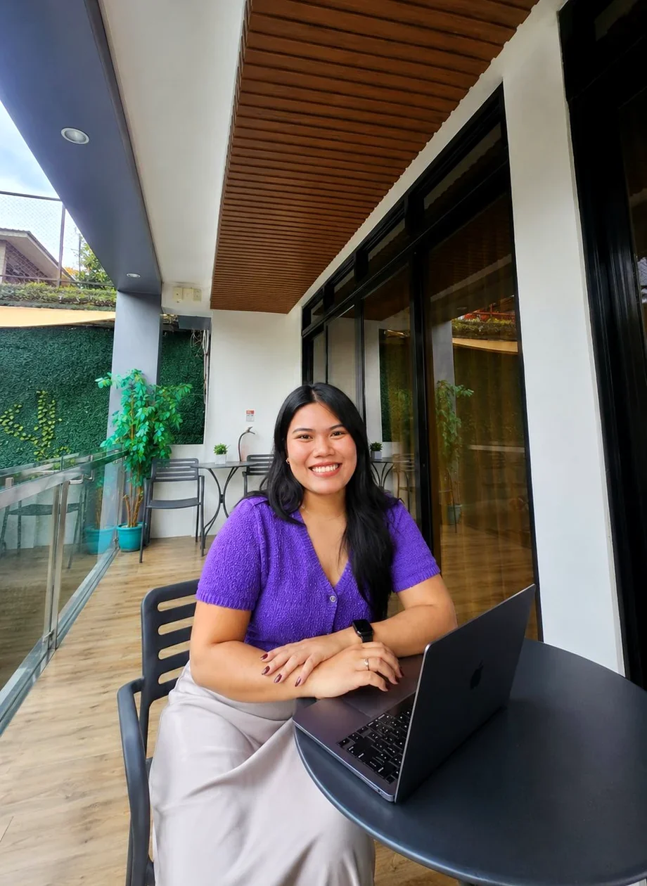 A woman with long black hair, wearing a purple top and beige skirt, sitting at a black table with a laptop on it, smiling indoors with large windows and wooden ceiling panels.