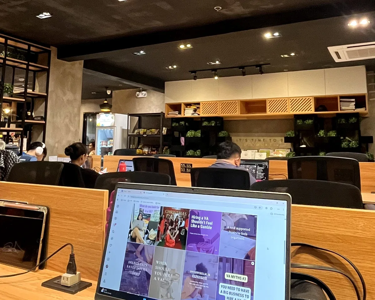 People working on laptops in a modern coffee shop or coworking space with wooden decor, shelves with greenery, and a refrigerated display in the background.