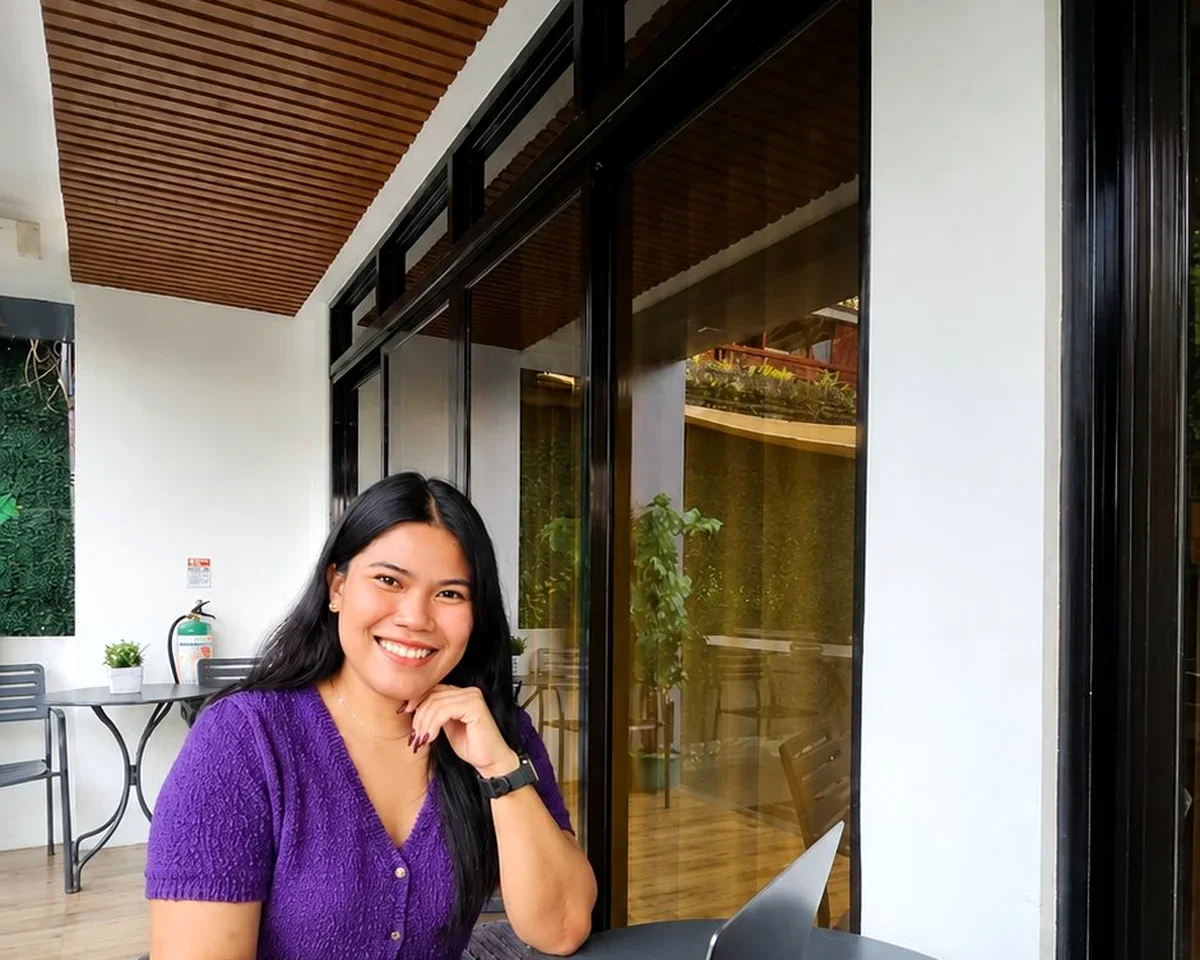 A woman with long black hair, wearing a purple blouse and a black watch, smiling and sitting at a table inside a modern cafe with large glass windows and wooden ceiling.