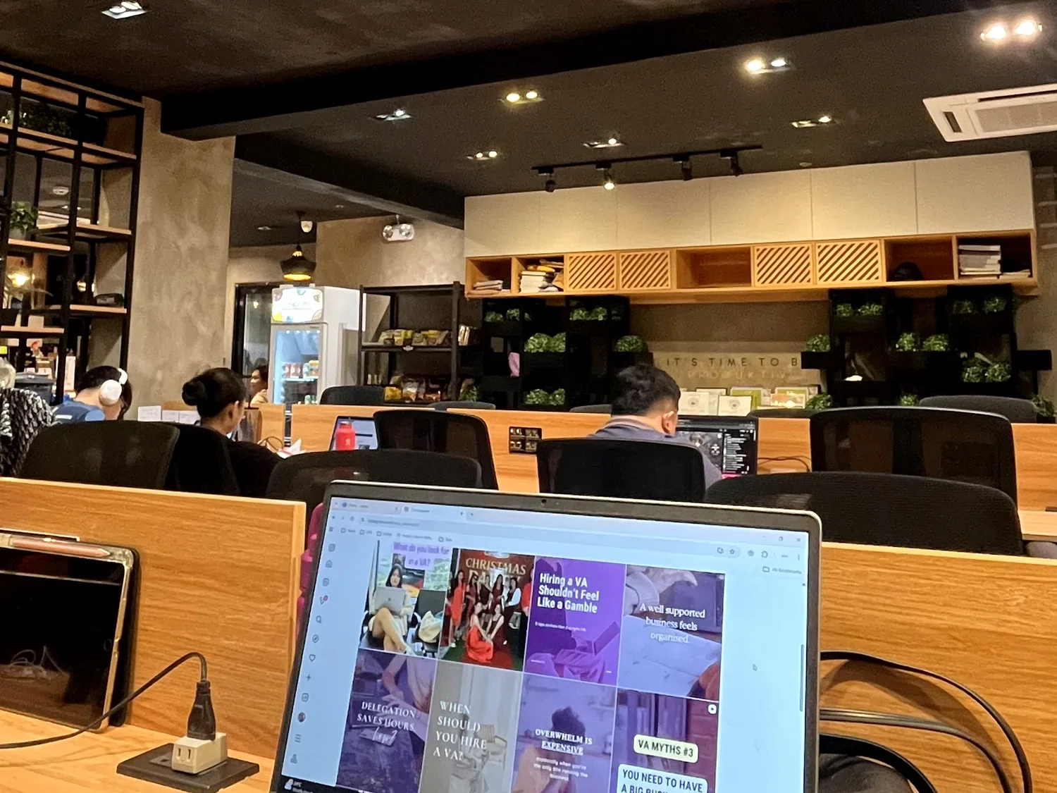 People working on laptops at desks in a cafe or coworking space with shelves, plants, and a refrigerator in the background.