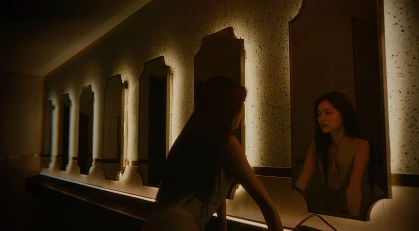 Une femme regarde dans un miroir éclairé, entourée de plusieurs autres miroirs sur un mur.