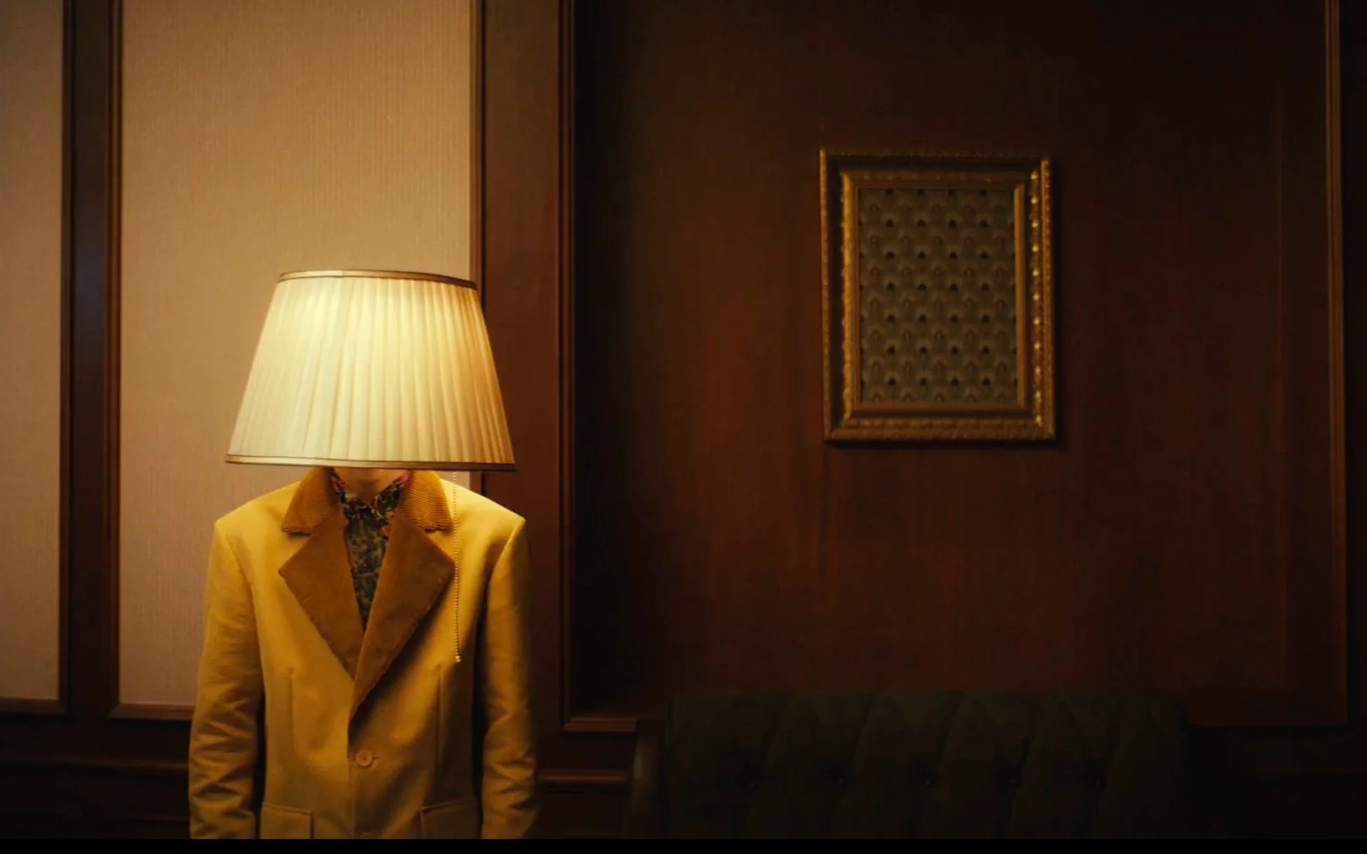 Personne avec un abat-jour comme tête, portant un manteau jaune, dans une pièce avec des murs en bois et un cadre vide.