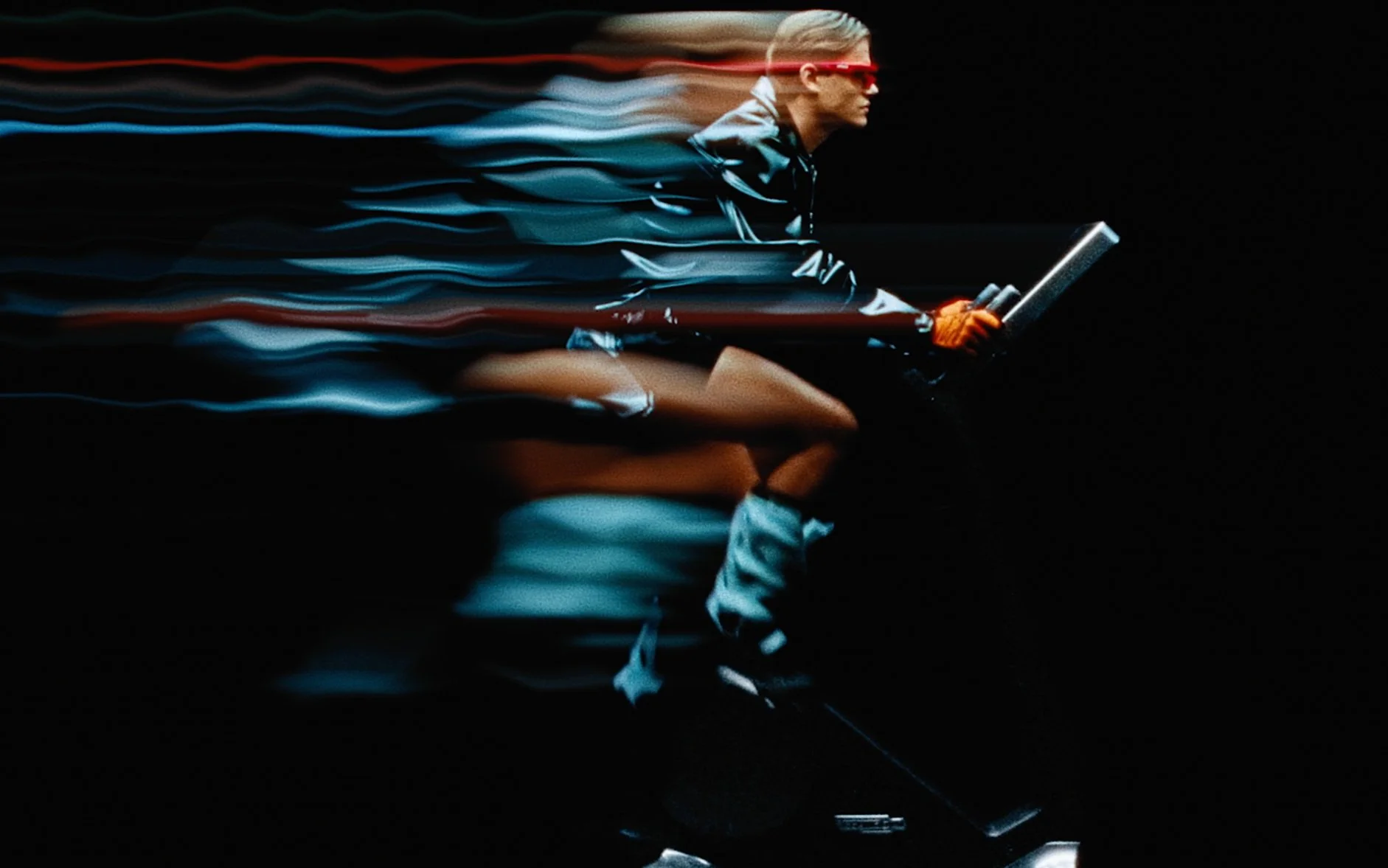 Une personne en mouvement rapide, portant des lunettes de soleil rouges et des gants orange, chevauchant un véhicule de deux roues avec des effets de flou lumineux.